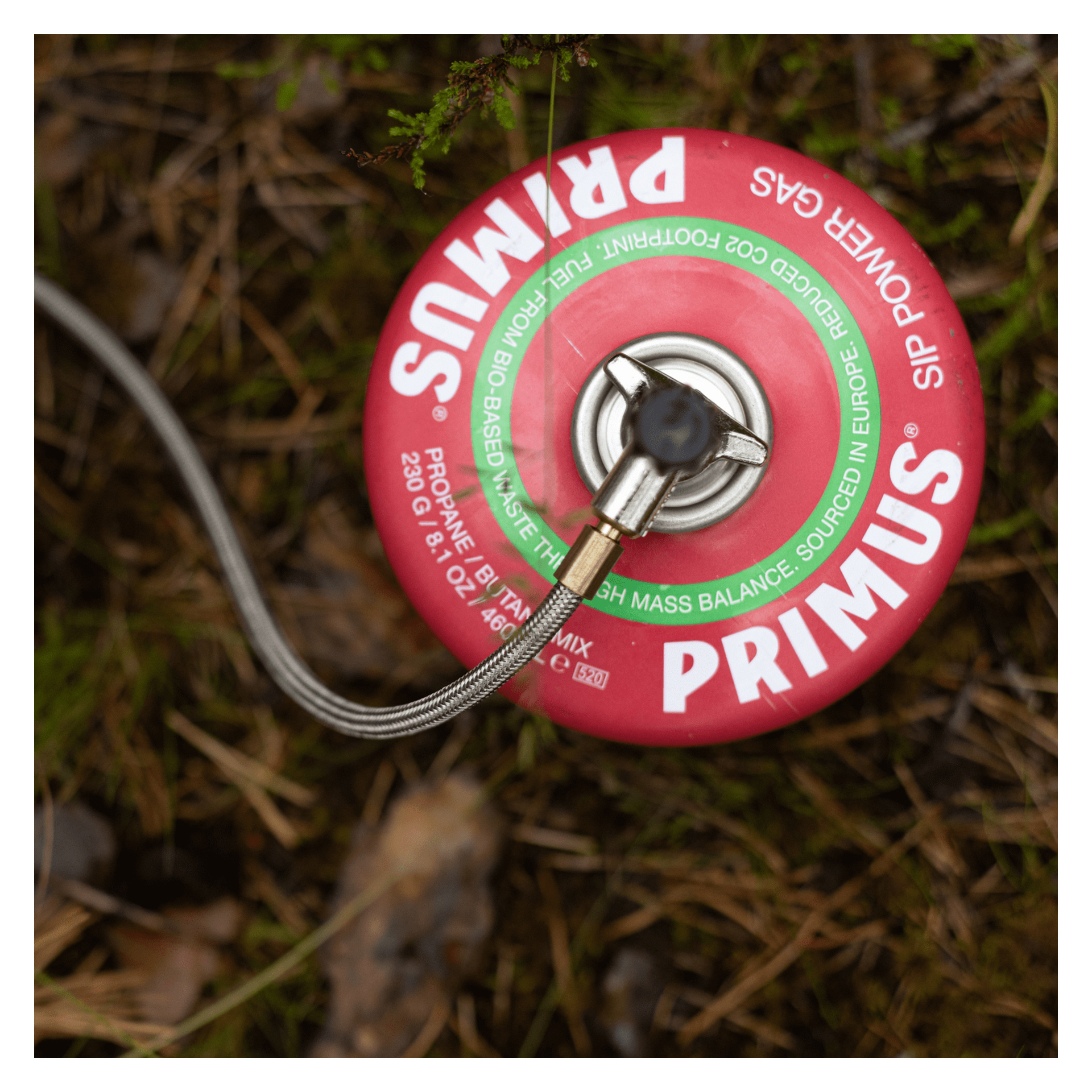 En röd Primus-propanbehållare med bränsleslang vilar på mossiga gräs och har etiketter med användnings- och säkerhetsdetaljer.