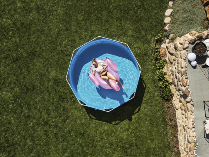 Von oben betrachtet entspannt sich eine Person auf einem rosa Schwimmer in einem blauen Becken, das auf Gras neben einer Steinmauer und einer Terrasse steht.