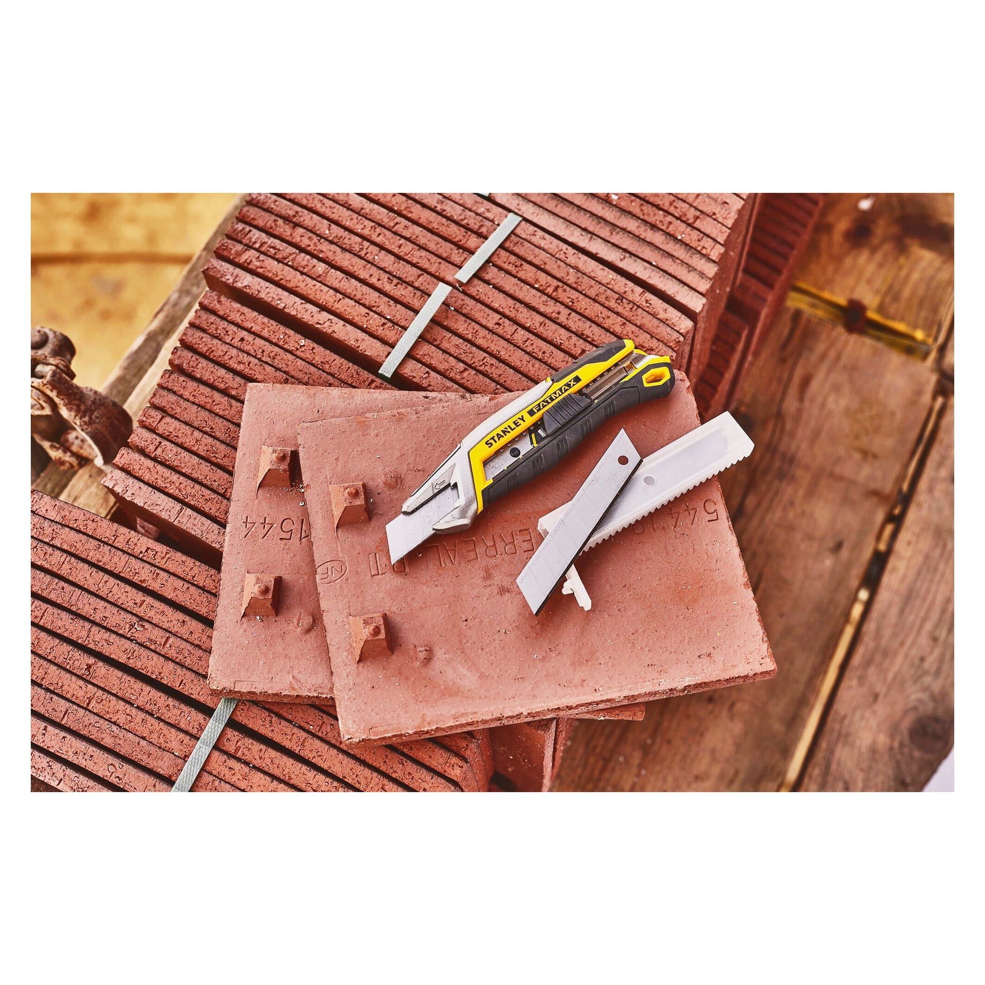 En gul-svart hobbykniv och ett silverblad som kan brytas av ligger på räfflade, rödbruna plattor ovanpå trä. En metallstång korsar plattorna.