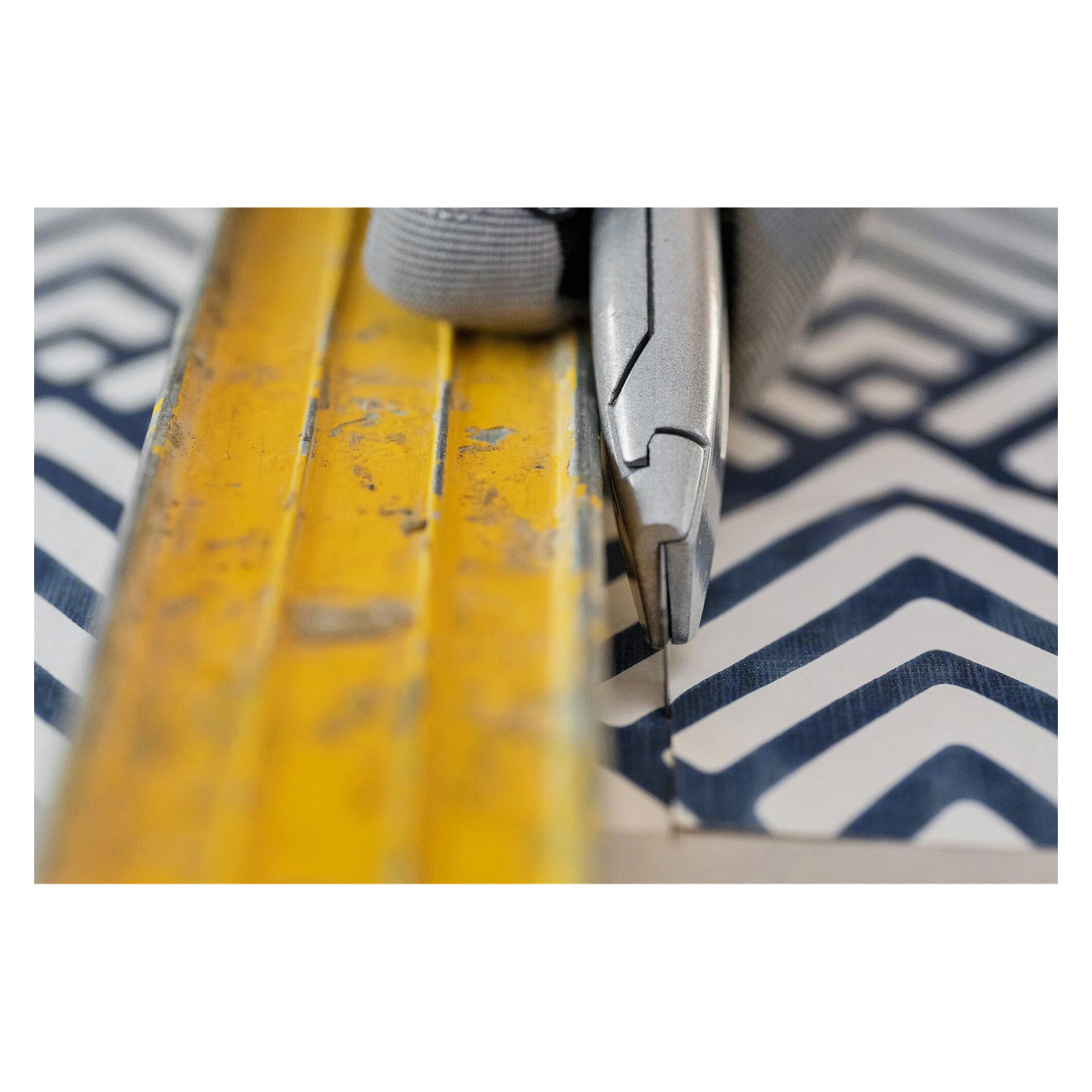 En handskbeklädd hand använder en hobbykniv och en gul metalllinjal för att skära blått och vitt mönstrat papper med geometriskt mönster.