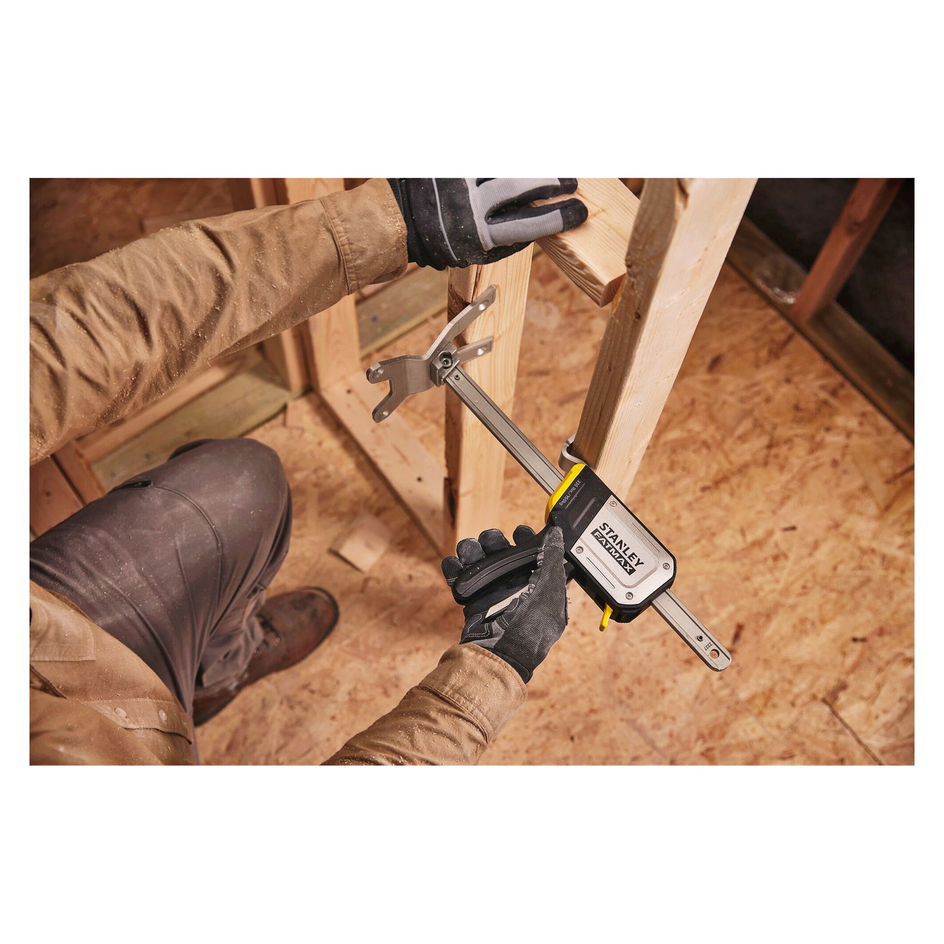 Med arbetshandskar fäster en person två träplankor på ett plywoodgolv med en Stanley FATMAX-klämma på en byggarbetsplats.