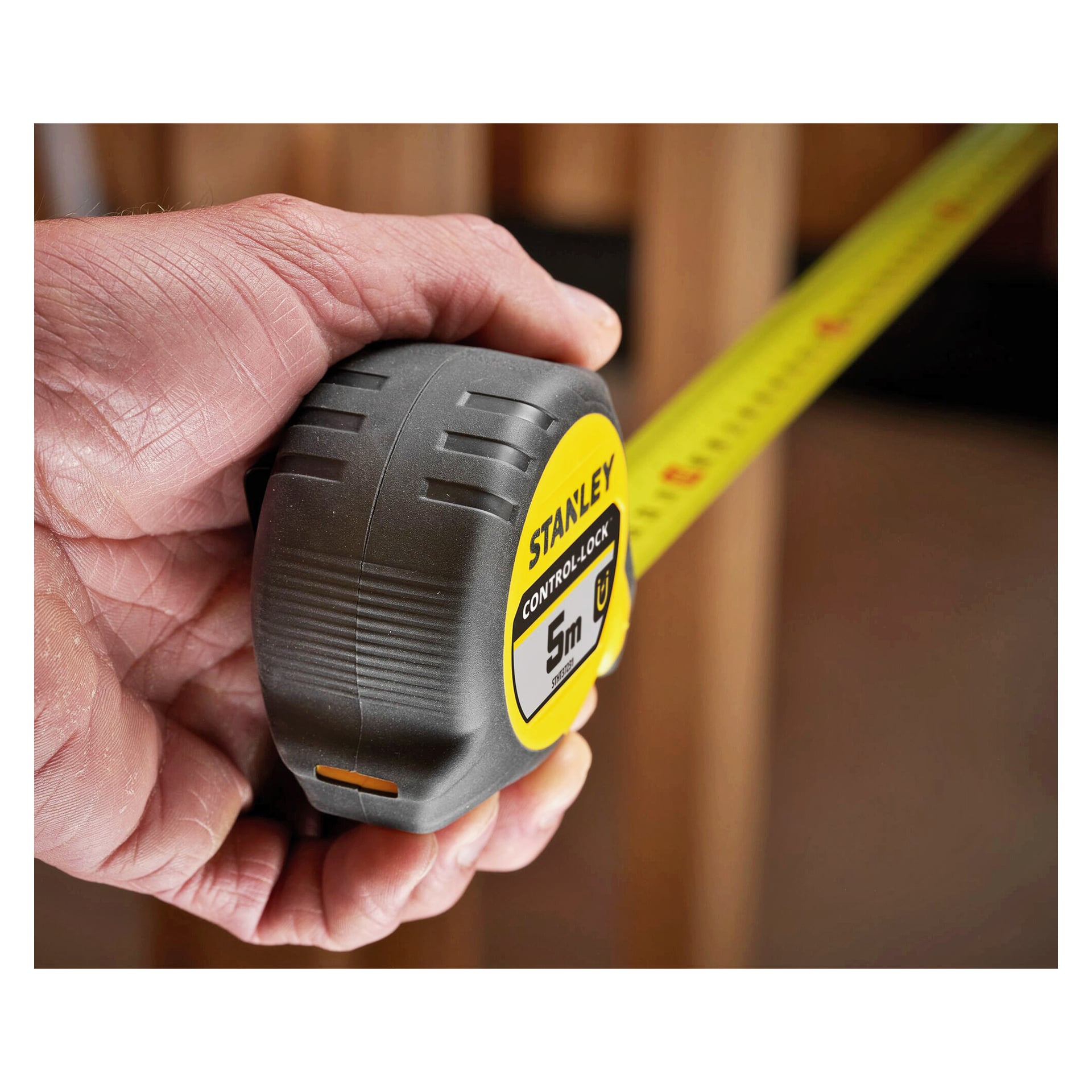 En hand håller ett Stanley 5-meter måttband med utsträckt gult band som visar siffror, placerat mot träbalkar i bakgrunden.