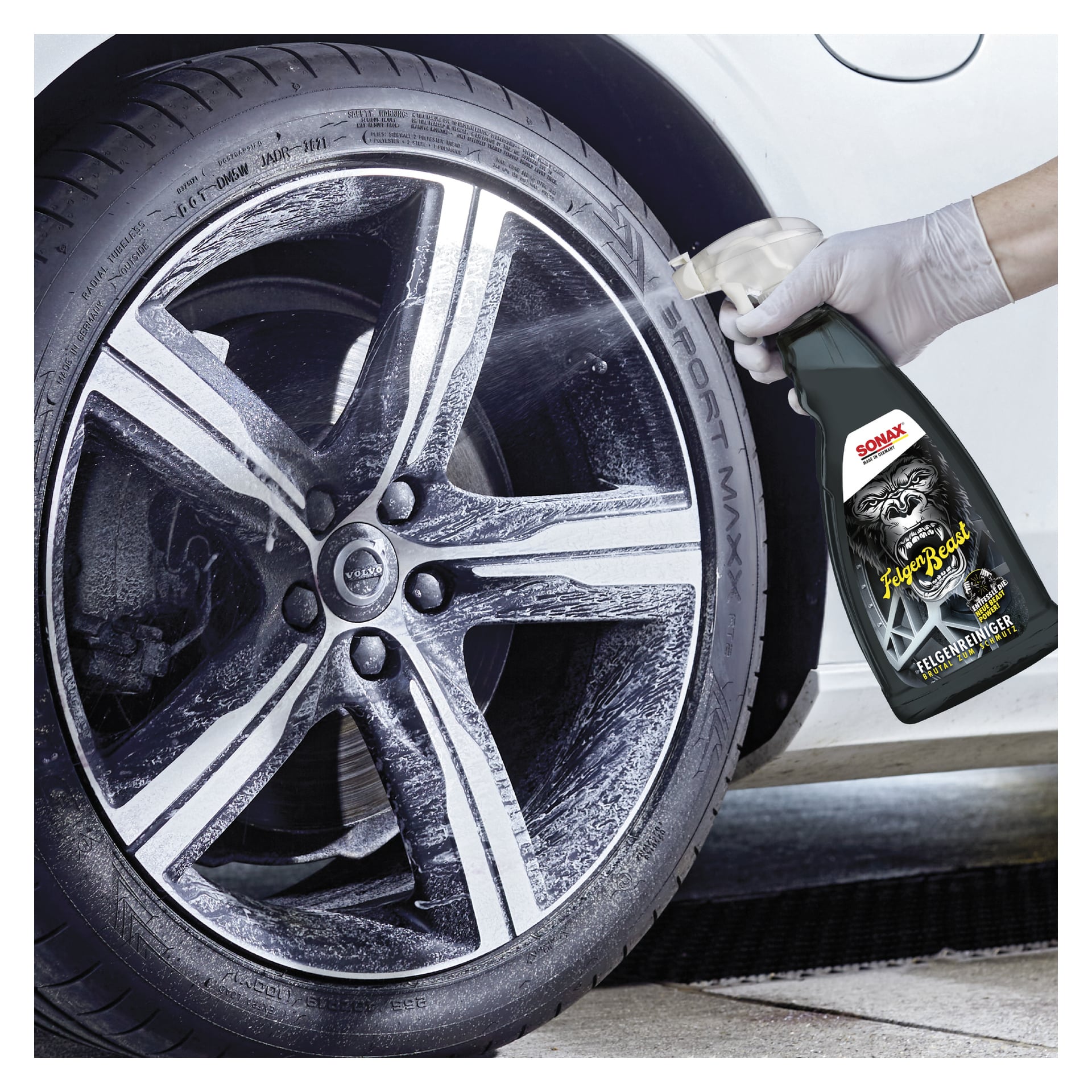 En handskbeklädd hand sprayar SONAX Xtreme Rim Cleaner på en lättmetallfälg, delvis skummat. SPORT MAXX är på däcket bredvid en vit bil utomhus.