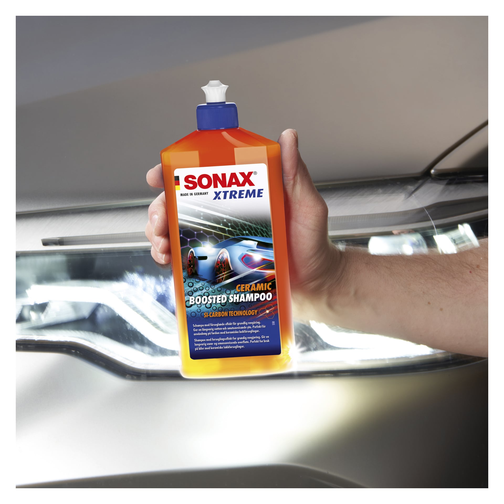 En hand håller en Sonax Xtreme Ceramic Boosted Shampoo-flaska nära en bilstrålkastare, med etiketten vänd mot betraktaren.