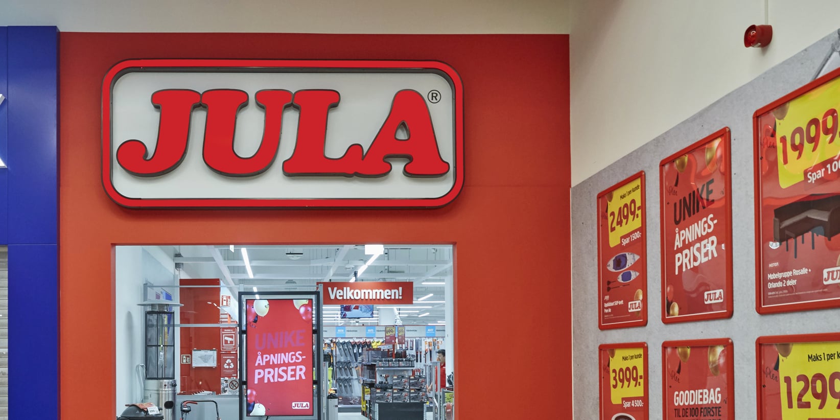 Inngangen til Jula-butikken med et stort rødt og hvitt Jula-skilt, pris- og kampanjeplakater på veggen, og produkter som kan ses inne i den lyse butikken.