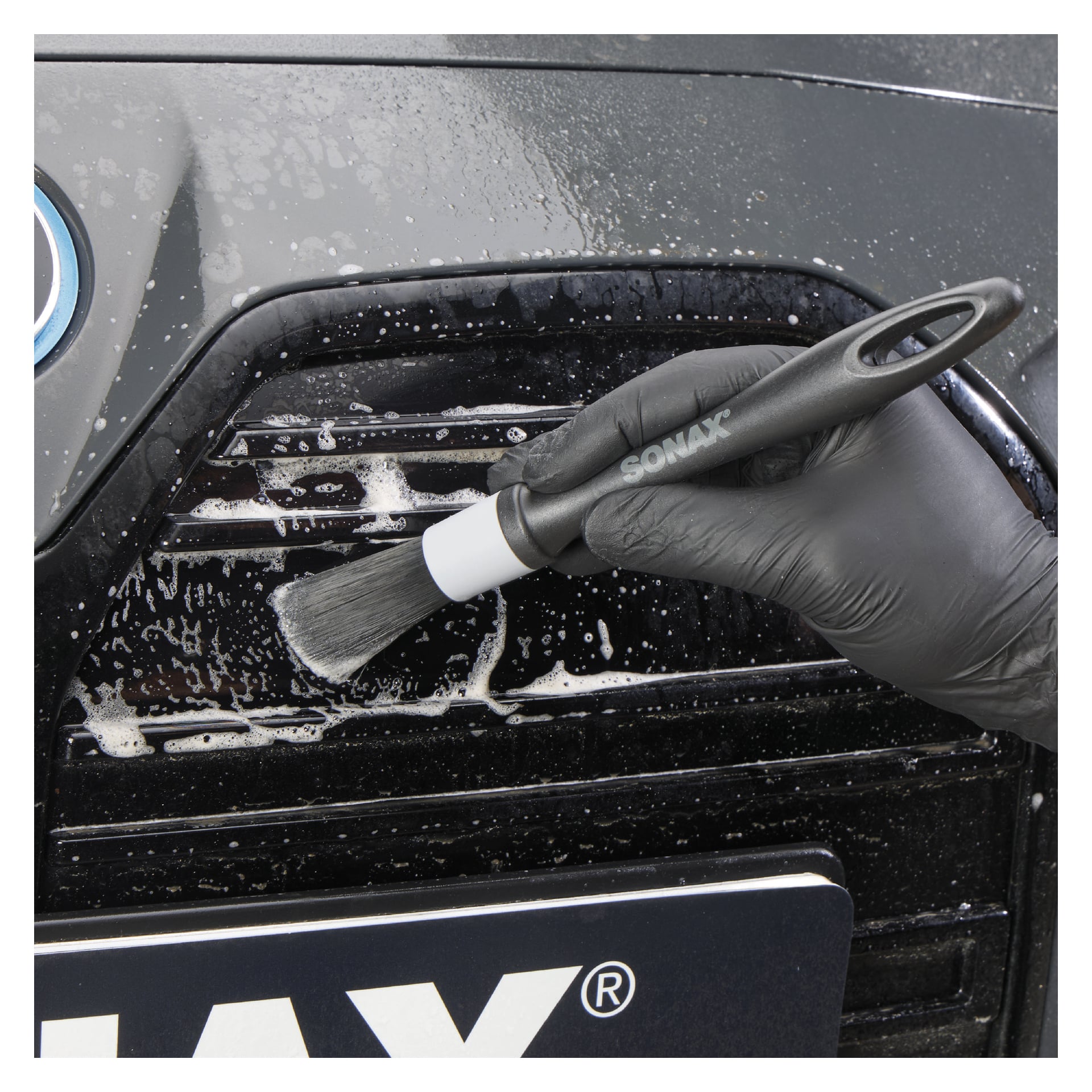 En hand med handske använder ett Sonax Deep cleaning Brushes Profiline 2-pack för att ta bort tvåliga rester från bilens svarta grill.