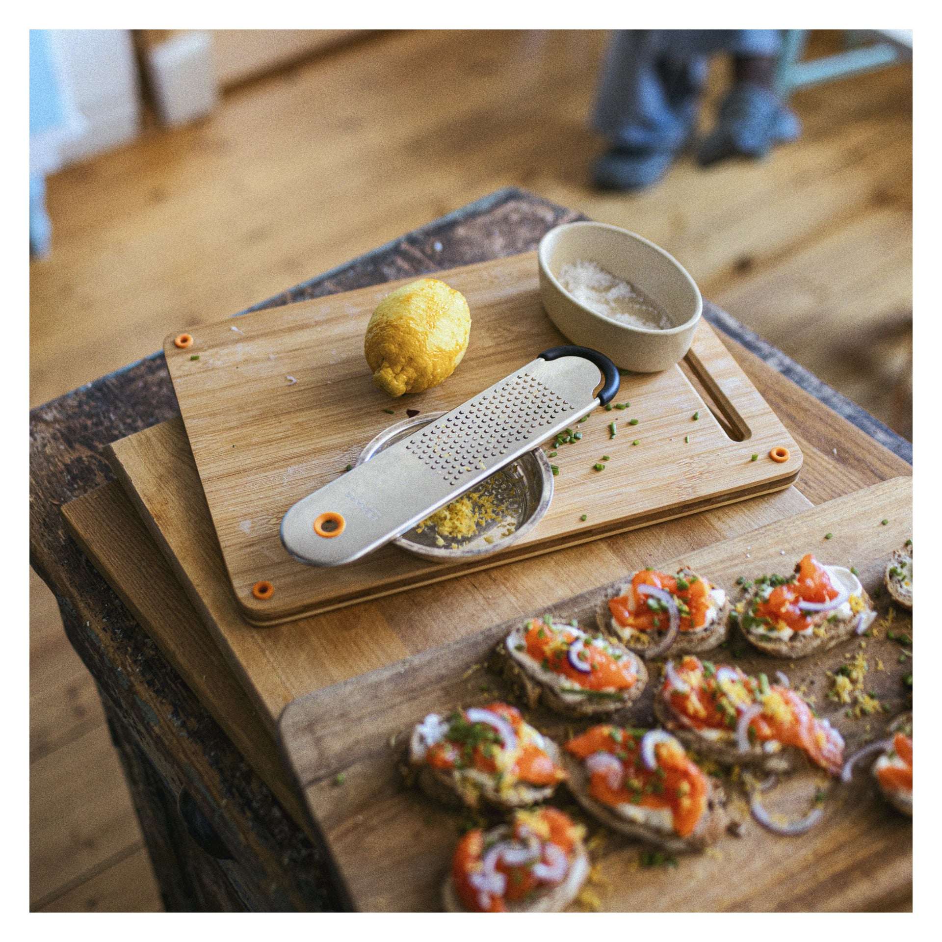 Na rustykalnym stole leży drewniana deska do krojenia z formą Fiskars Extra Fine Grater Functional Form, cytryna, miska wypełniona drobno startą skórką oraz kilka kawałków chleba z łososiem, cebulą i ziołami. W rozmytym tle nogi są częściowo widoczne.