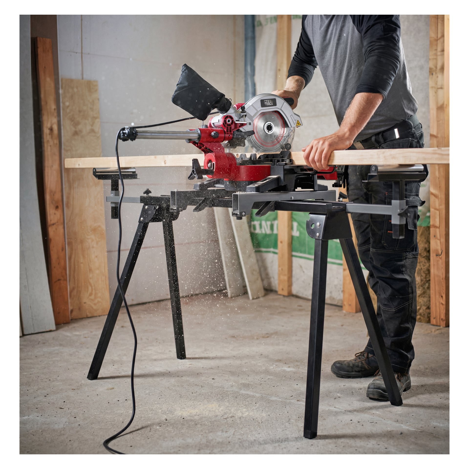 En person använder en röd elektrisk geringssåg monterad på ett Meec Tools Saw Stand, och sågar en träplanka i ett byggområde. Sågspån finns där, och omgivningen har trästomme och ofärdiga väggar.