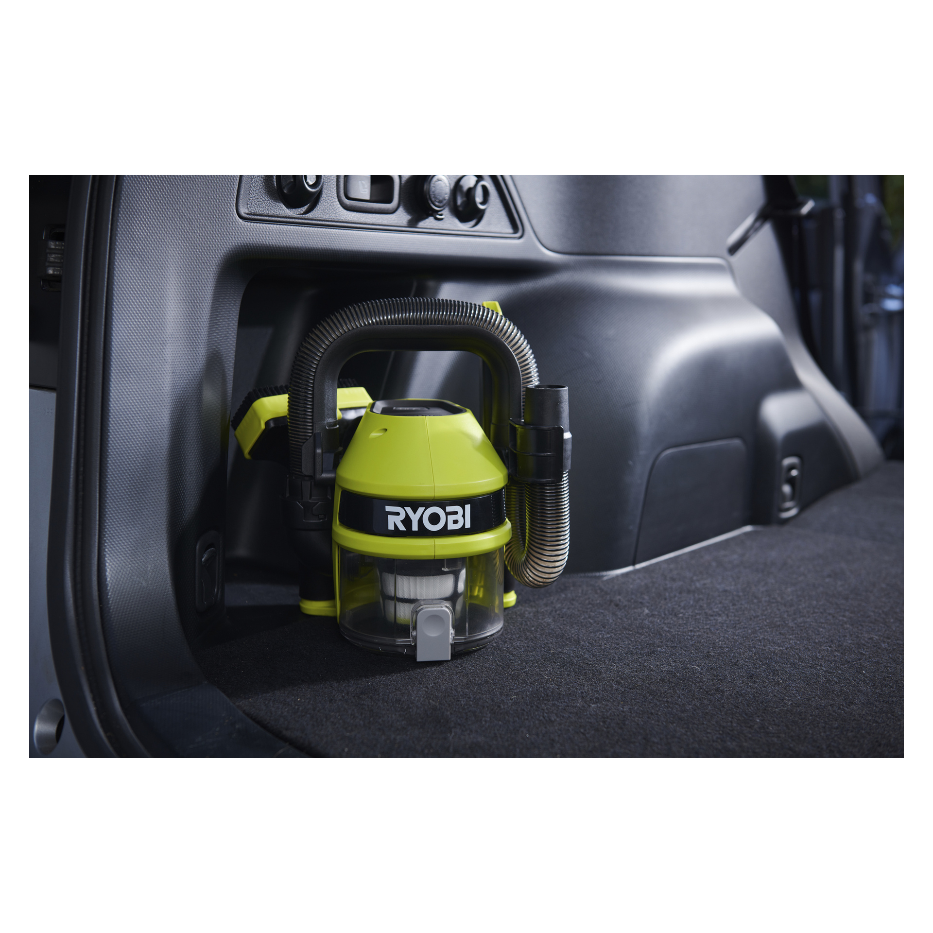 En kompakt Ryobi One+ batteridrevet bilstøvsuger (RAV 1805-0) er plassert på bilens mørke teppebelagte gulv, slangen pent viklet rundt karosseriet, mens et kontrollpanel vises i bakgrunnen.