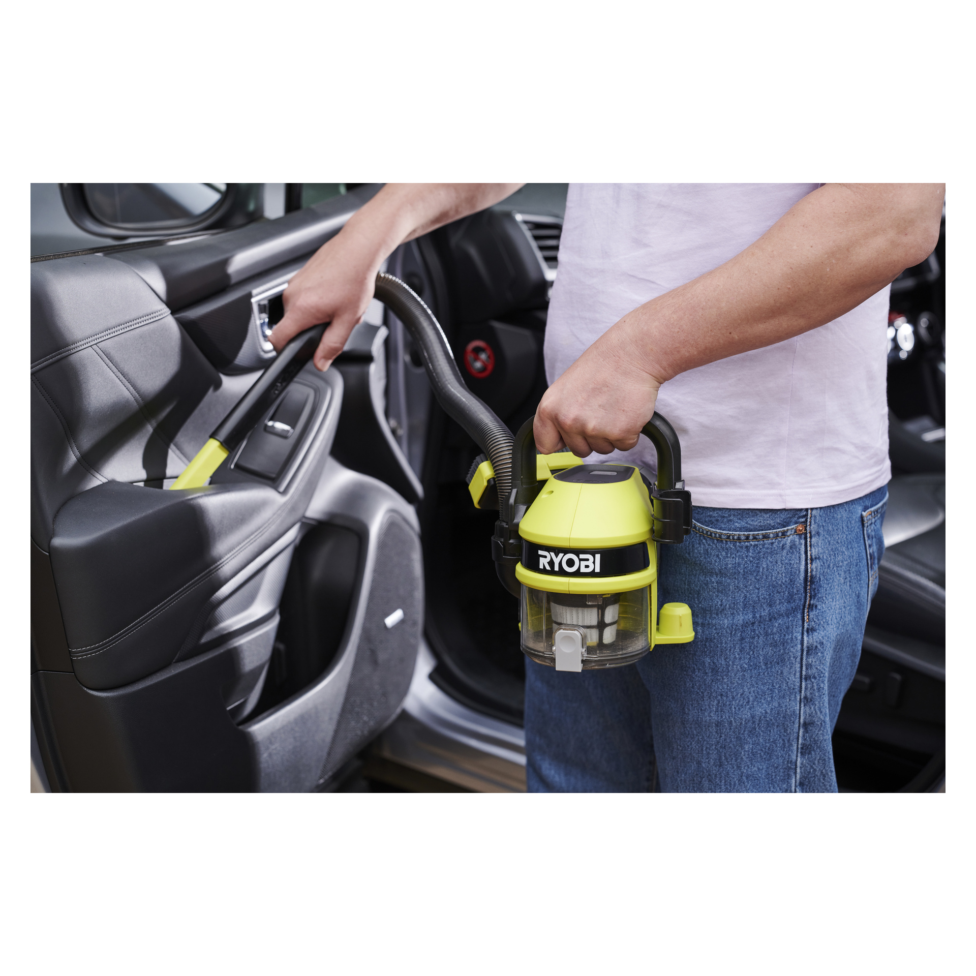 En person i jeans står utenfor en bil og holder Ryobi One+ batteridrevne bilstøvsuger RAV 1805-0. De bruker vakuumslangen til å rengjøre dørpanelet, og leder det inn i kjøretøyets interiør mens de støtter hovedbeholderen.