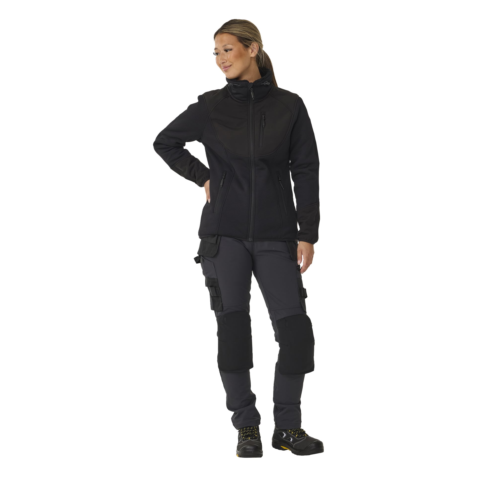 En kvinna modellerar Bluewear Women's Moa fleecejacka med luvfoder, stylad med mörkgrå cargobyxor med knäskydd och svart-gula arbetsskor. Hennes långa hår är uppsatt och hon tittar åt sidan, ena handen i fickan.