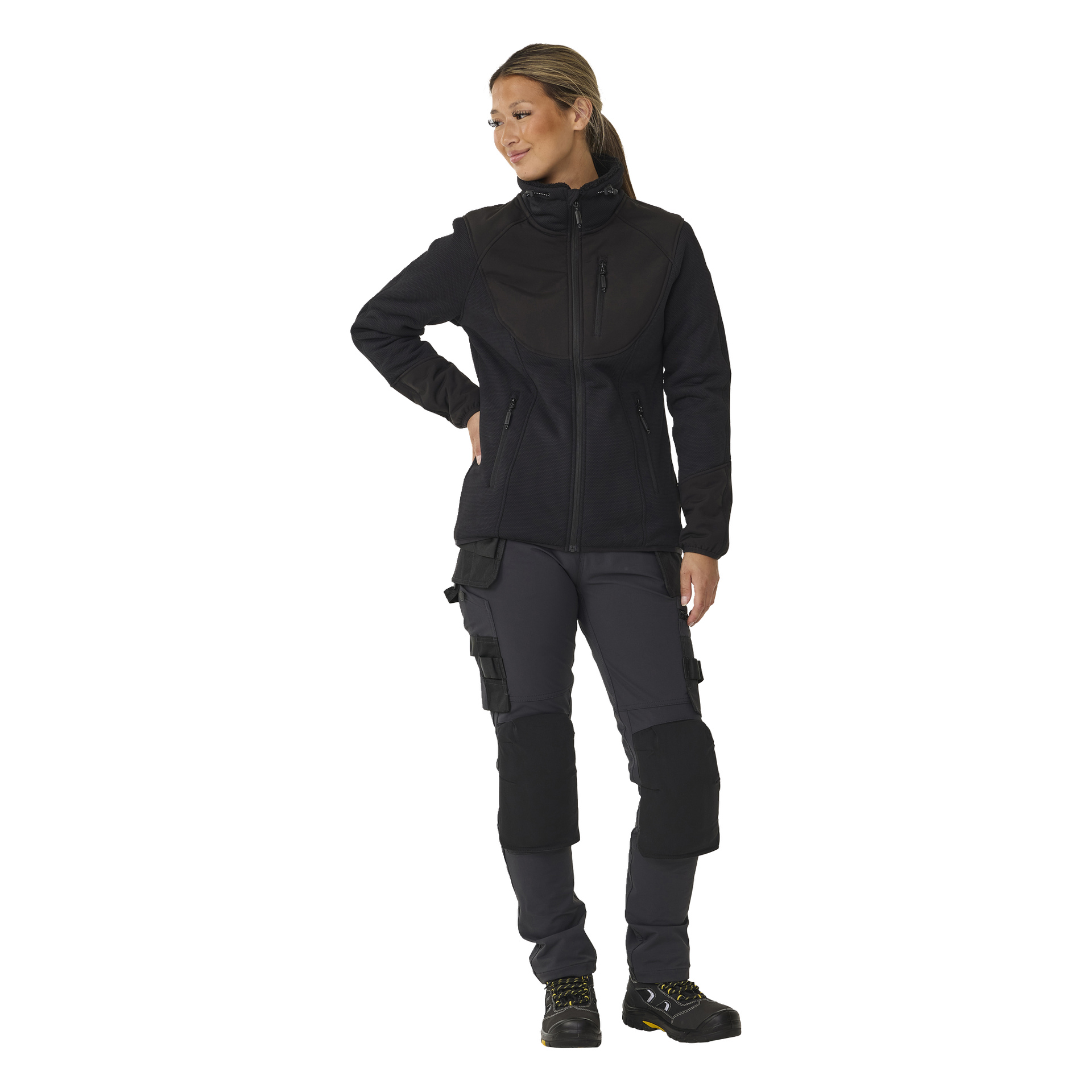 En kvinne modellerer Bluewear Women's Moa-fleecejakke med luvfôr, stående med den ene hånden på hoften. Hun har håret i hestehale og er kledd i mørke cargobukser med knebeskyttere, samt svarte og gule joggesko. Hun ser til siden med et svakt smil.