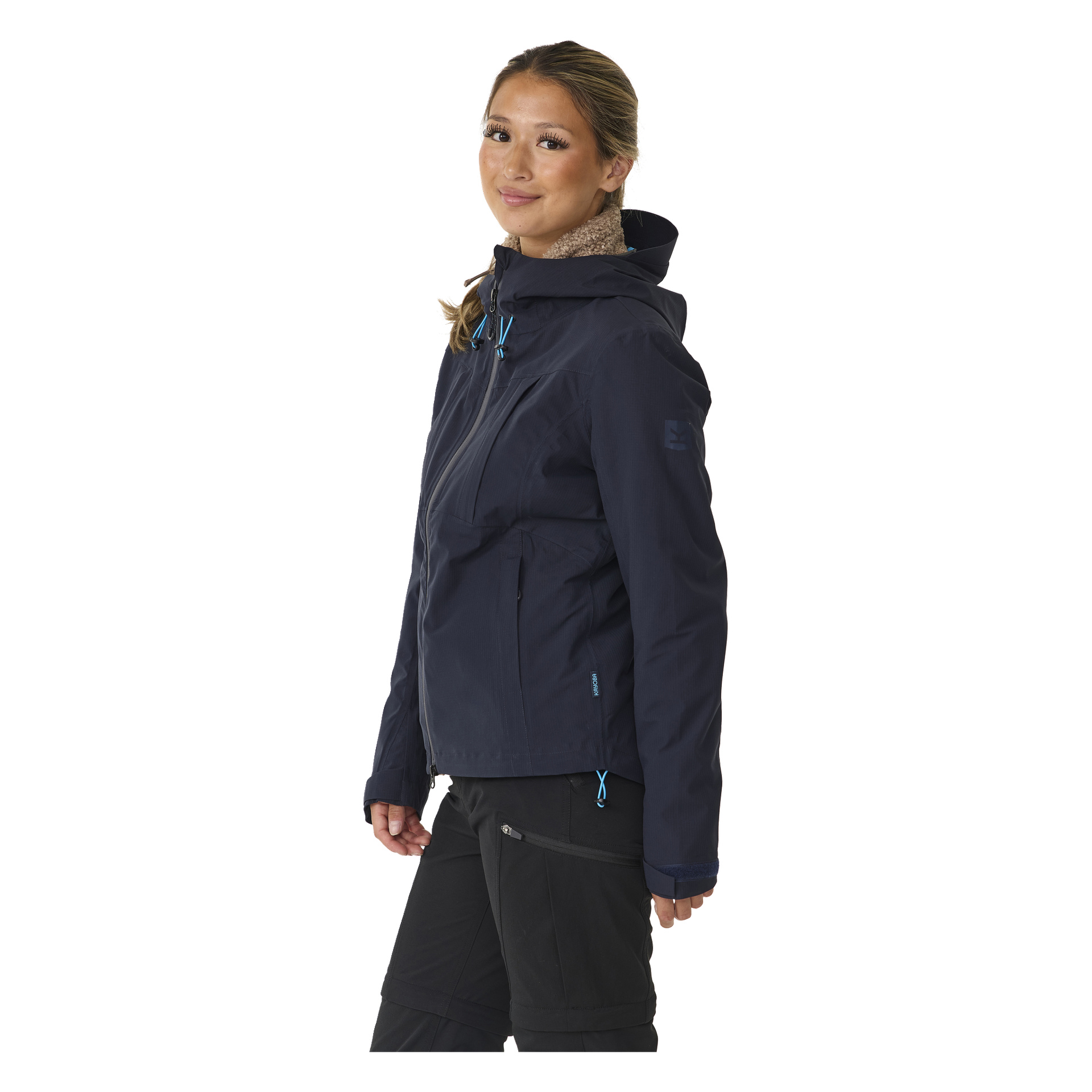 Eine Person steht leicht gedreht, trägt eine marineblaue Kayoba Outdoor Damenjacke mit blauen Reißverschlüssen und schwarze Hosen. Ihr Haar ist zurückgebunden und ihr Gesichtsausdruck neutral.