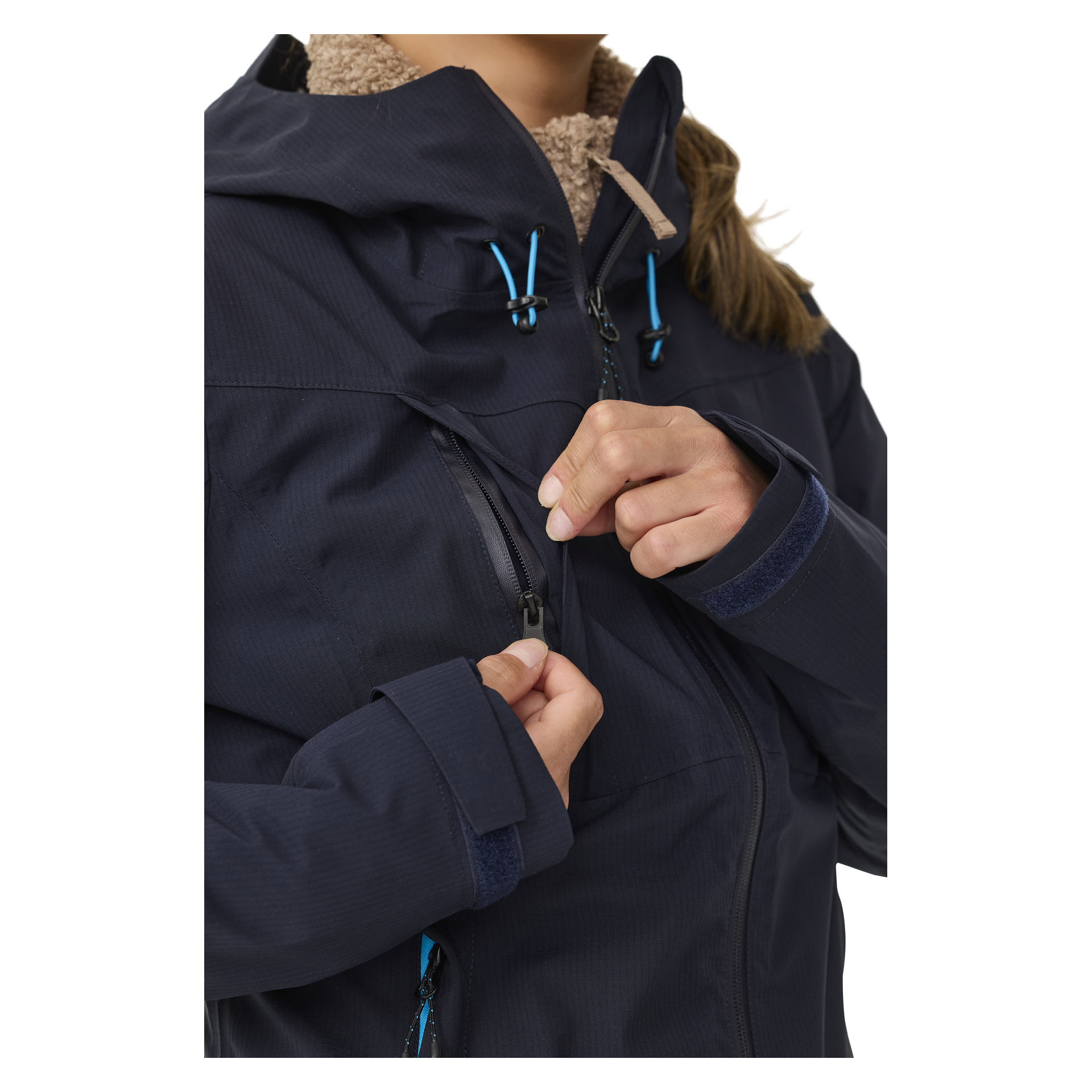 Eine Person, sichtbar vom Oberkörper aufwärts, trägt die Kayoba Outdoor Women's Shell-Jacke Sarek. Die Jacke ist dunkel mit blauen Kordeln und Hakenschlaufen an den Handgelenken. Die Person ist gerade dabei, eine Brusttasche zu öffnen.