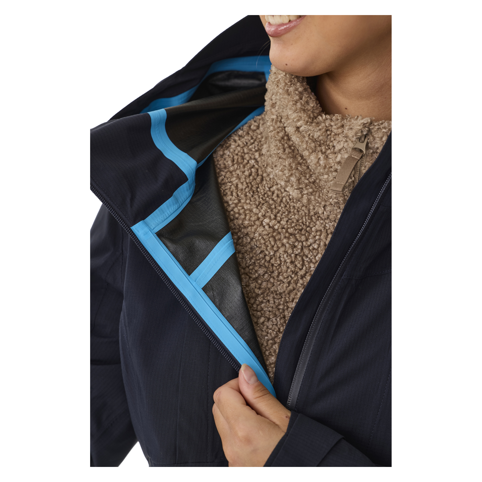 Nur die untere Hälfte des Gesichts einer lächelnden Person ist sichtbar, da sie einen beigefarbenen, strukturierten Fleecepullover unter einer teilweise geöffneten Kayoba Outdoor Women's Sarek Shell-Jacke trägt. Die Jacke hat eine dunkle Außenseite mit blauen Nähten, und das Innenfutter ist freiliegend.