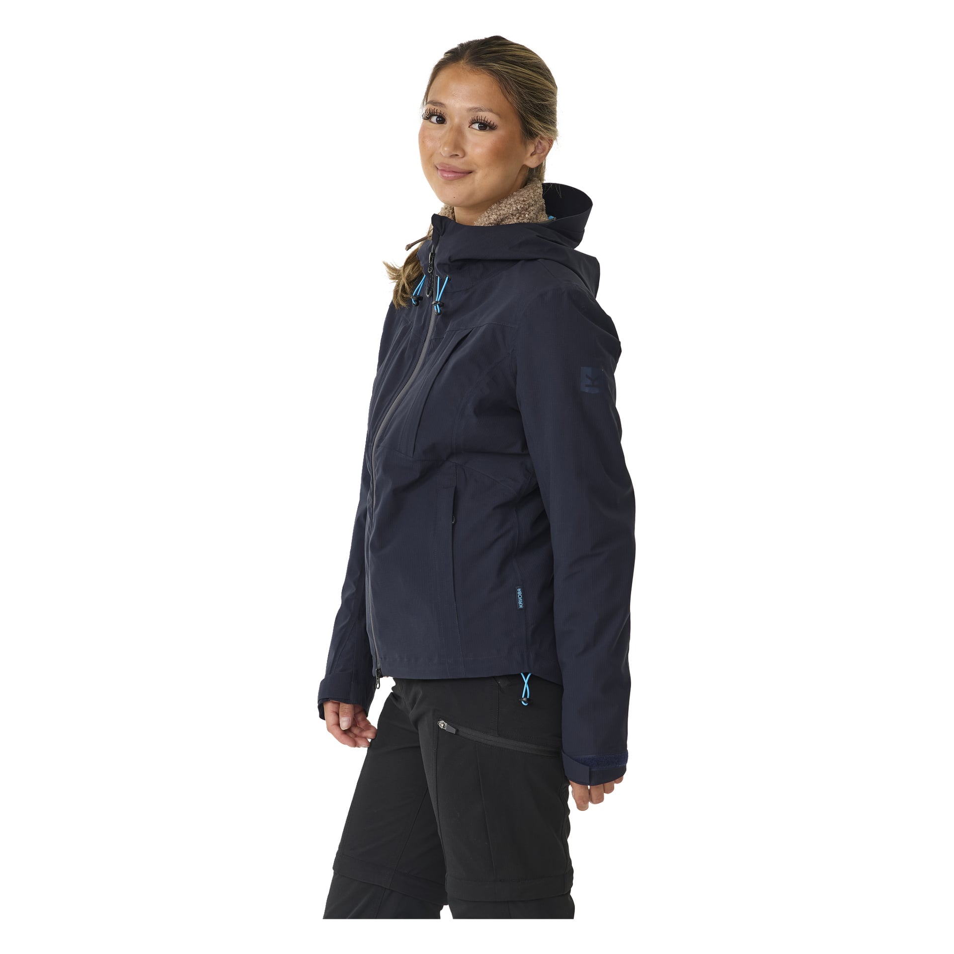Eine Frau steht leicht nach links gedreht, lächelt mit zurückgebundenem Haar. Sie trägt eine dunkelblaue Kayoba Outdoor Damen-Shelljacke Sarek mit blauen Reißverschlüssen und hohem Kragen, kombiniert mit schwarzen Hosen.