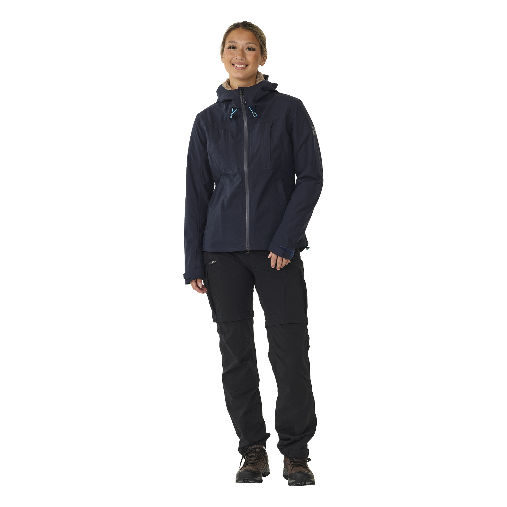 Eine lächelnde Person steht nach vorne gewandt, gekleidet in die Kayoba Outdoor Women's Shell Jacket Sarek, schwarze Hosen und braune Wanderschuhe, mit entspannten Armen an den Seiten.