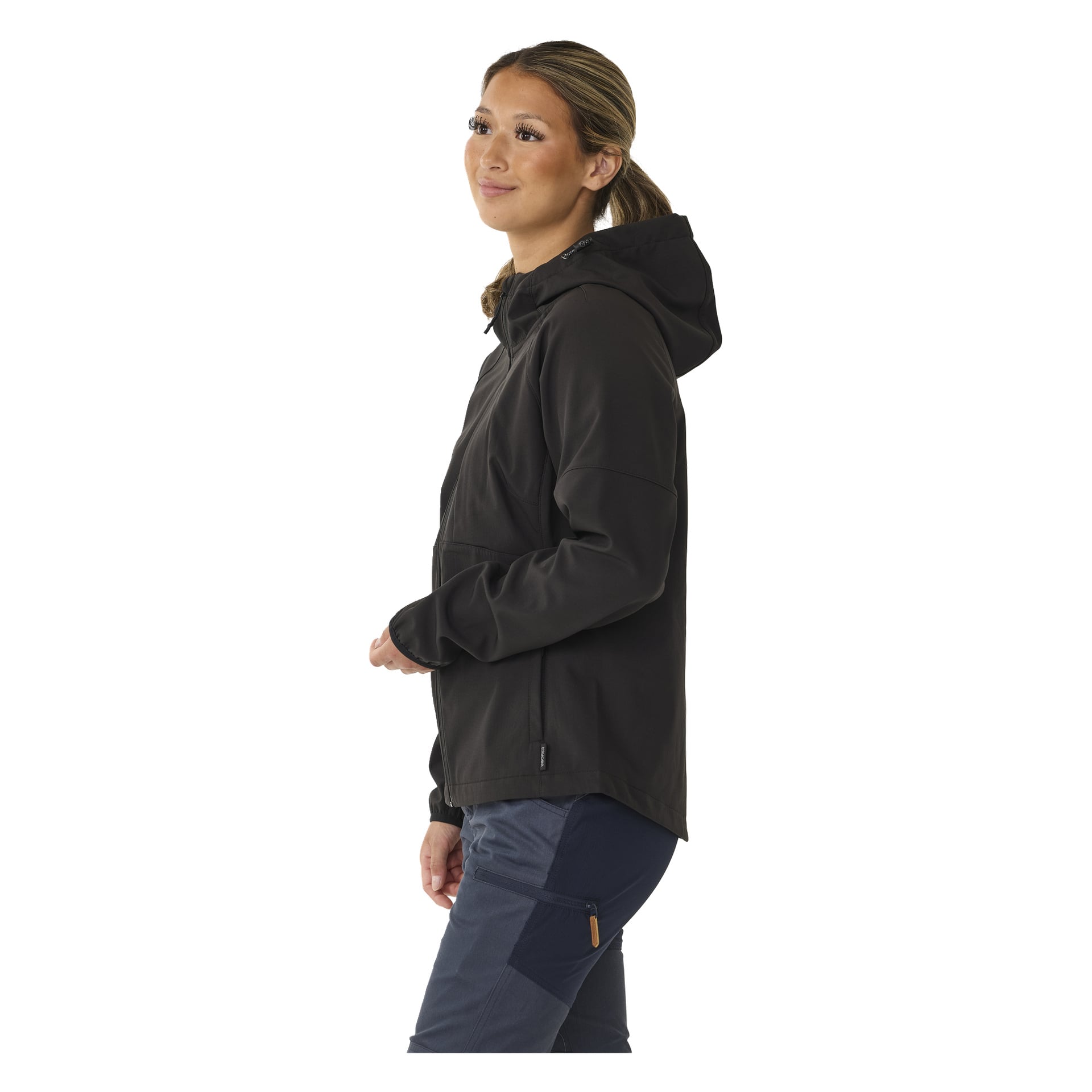 Eine Person mit langen, zurückgebundenen Haaren steht leicht zur Seite geneigt, gekleidet in die mit Reißverschluss verzauste Kayoba Outdoor Softshell-Softshell-Jacke Hede, die in Schwarz mit einer mit Reißverschluss gefüttert wurde, kombiniert mit marineblauer Hose.