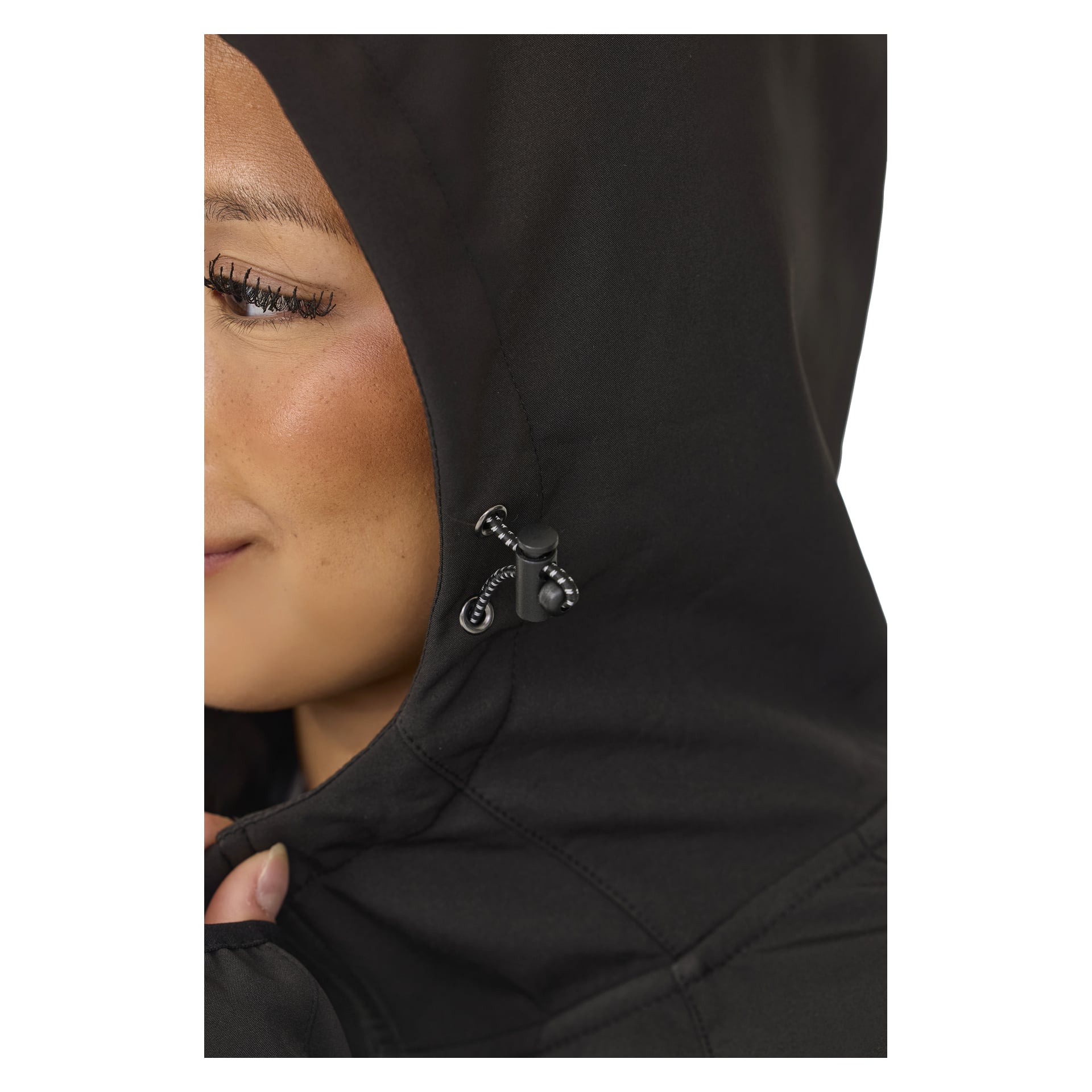 Eine Nahaufnahme zeigt eine Person in der Kayoba Outdoor Women's Softshell-Jacke Hede, die die Seite des Gesichts hervorhebt, sowie den verstellbaren Kordelzug-Schalter an der Kapuze. Sie haben ein sanftes Lächeln und greifen leicht an den Rand der Kapuze.
