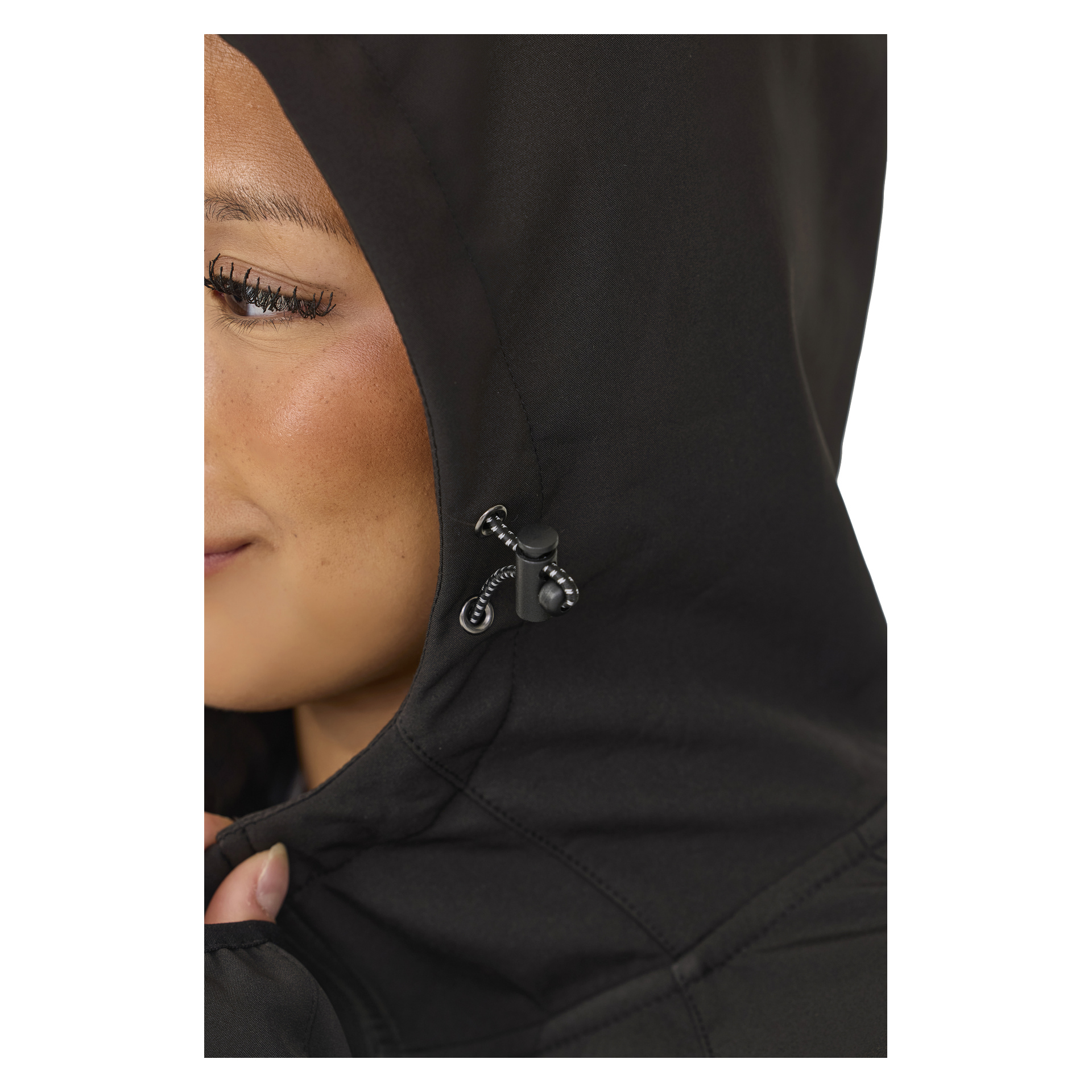 Eine Nahaufnahme zeigt jemanden, der die Kapuze einer schwarzen Kayoba Outdoor Women's Softshell-Jacke Hede mit Fleecefutter richtet, wobei der Fokus auf dem markanten schwarzen Kippschnurschloss liegt. Das Bild konzentriert sich auf die Gesichtszüge der Jacke, während nur ein Teil des Gesichts des Trägers sichtbar ist.