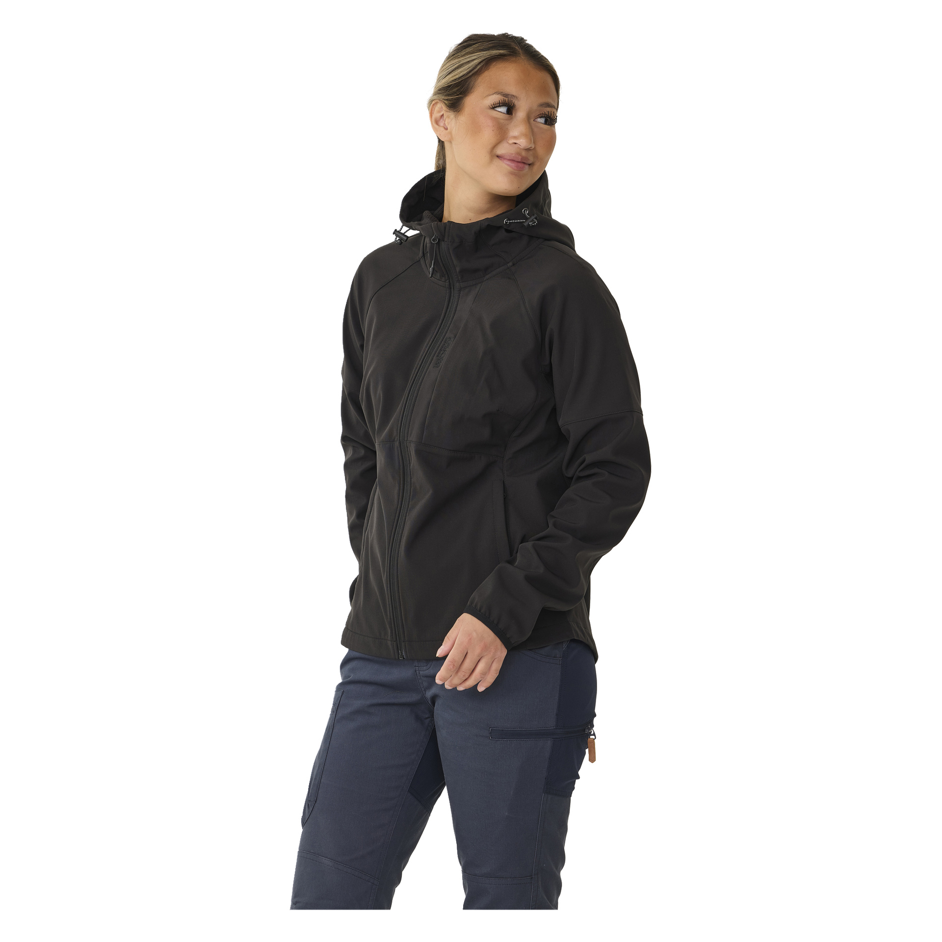 Eine Person mit hellem Haar, das nach hinten gebunden ist, trägt die Kayoba Outdoor Women's Softshell-Jacke Hede, die mit einer dunkelblauen Hose kombiniert wird. Sie stehen auf und blicken leicht nach rechts mit neutralem Gesichtsausdruck.