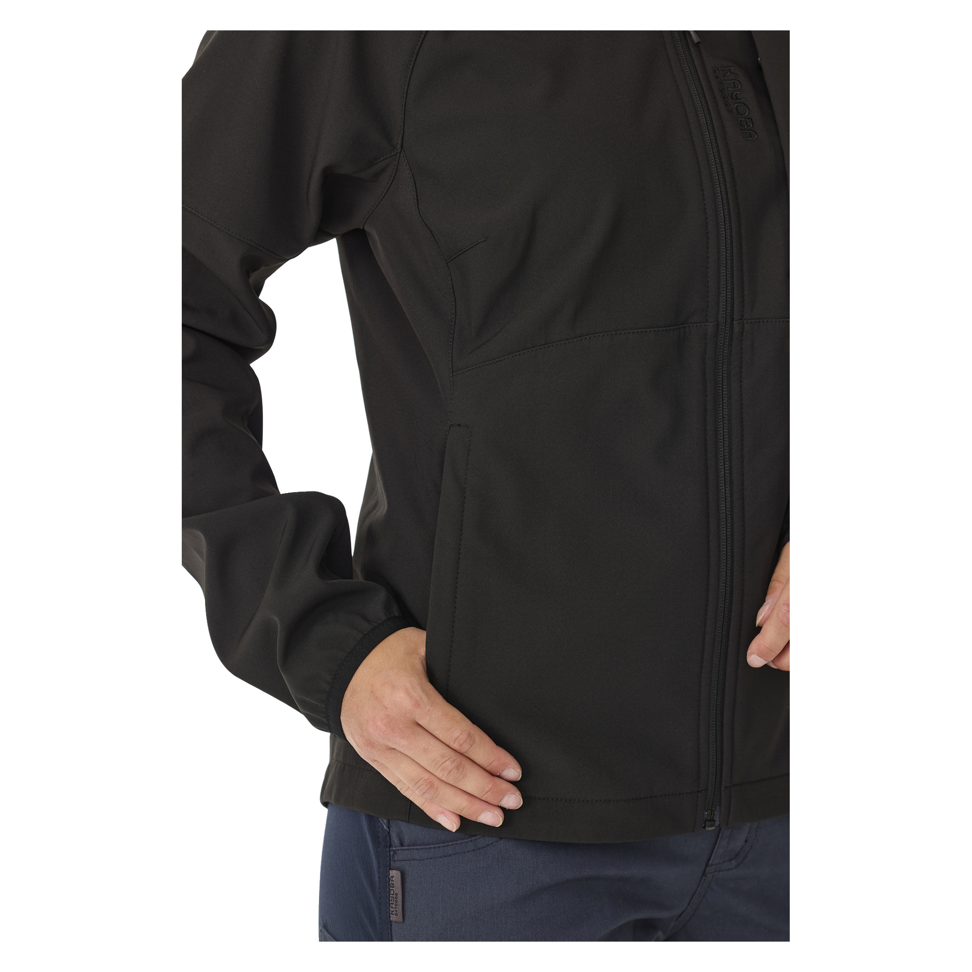 Eine Person ist von den Schultern bis zu den Hüften sichtbar, trägt eine schwarze, mit Fleece gefütterte Kayoba Outdoor Women's Softshell-Jacke Hede und dunkelblaue Hosen. Eine Hand befindet sich in der Nähe einer Tasche der Jacke, die andere in der Nähe des Reißverschlusses.
