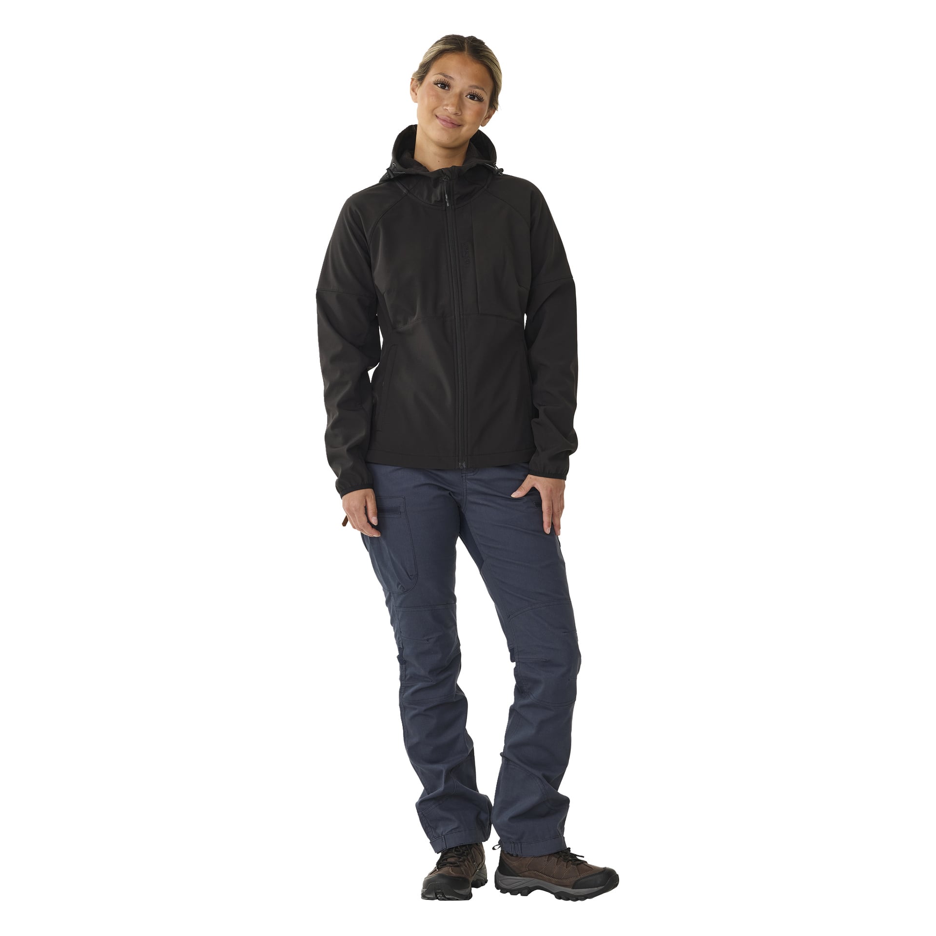 Eine Person steht mit dem Rücken nach vorne, gekleidet in die Kayoba Outdoor Women's Hede schwarze, fleecegefütterte Softshelljacke, blaue Cargohosen und braune Wanderschuhe. Ihr Kopf ist leicht geneigt und eine Hand ruht auf ihrem Oberschenkel.