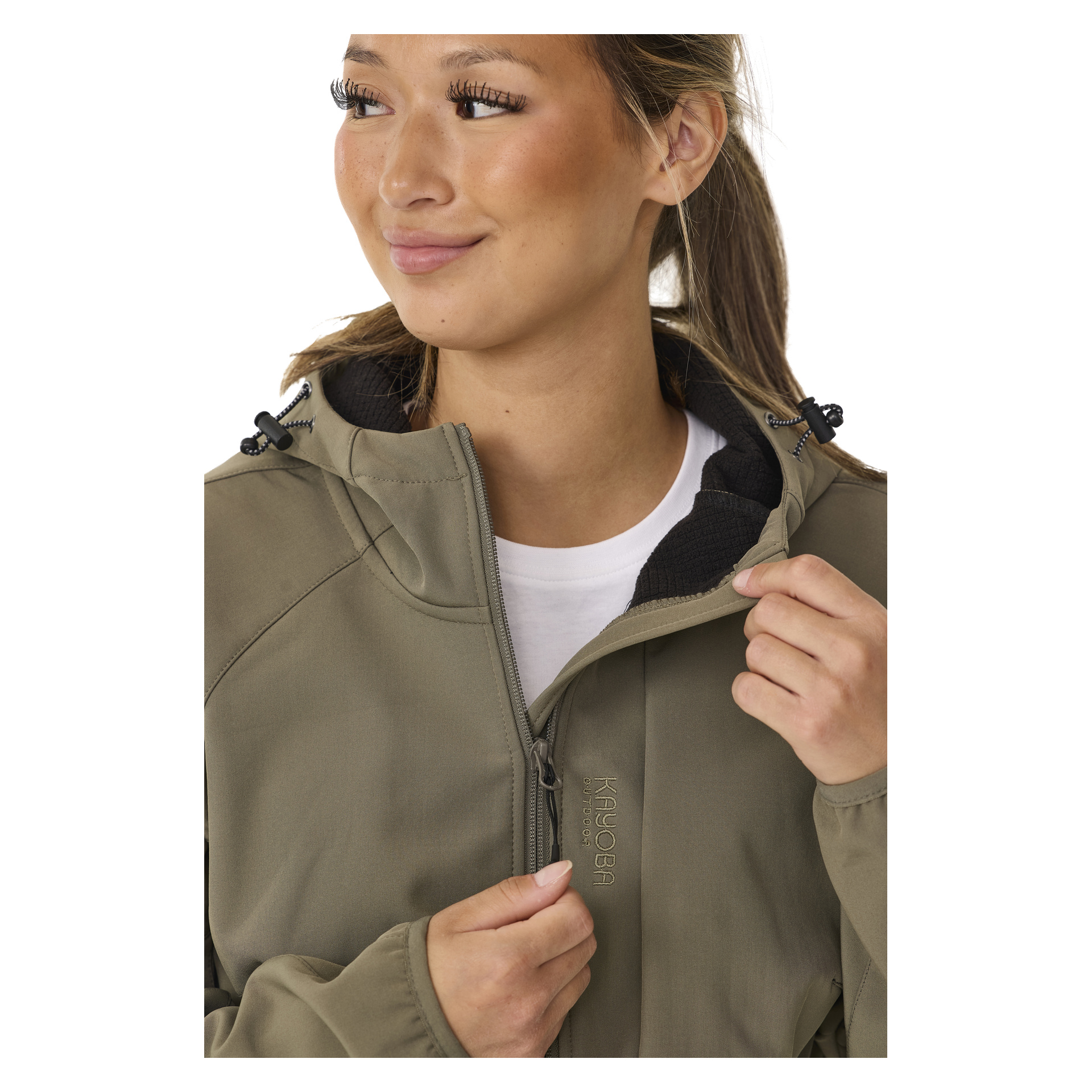 Eine lächelnde Person zieht eine helle, olivfarbene, mit Fleece gefütterte Damen-Softshelljacke zu, Hede von Kayoba Outdoor, mit dem Markennamen in der Nähe des Reißverschlusses. Sie tragen darunter ein weißes Hemd und schauen leicht zur Seite.