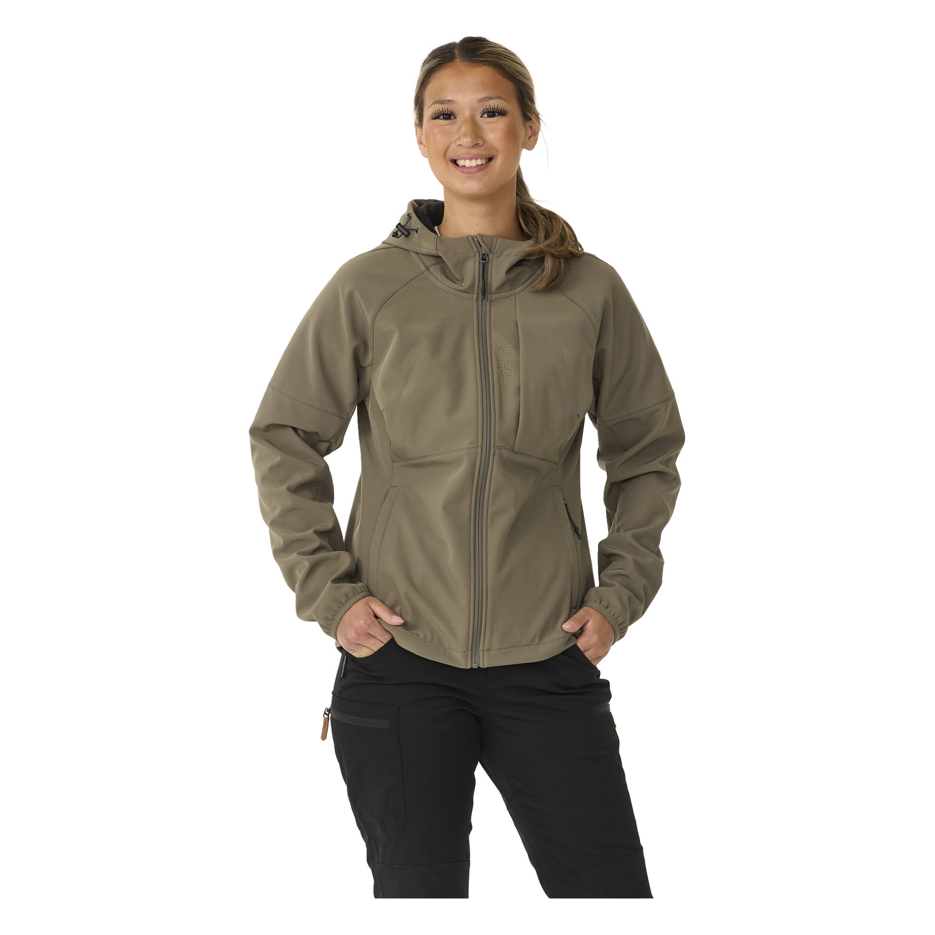Eine Person steht mit dem Rücken nach vorne, lächelt, gekleidet in die Kayoba Outdoor Women's Softshell-Jacke Hede, die mit Fleece gefüttert ist, in Khaki mit schwarzer Hose. Ihre Hände sind in den vorderen Taschen der Jacke versteckt.