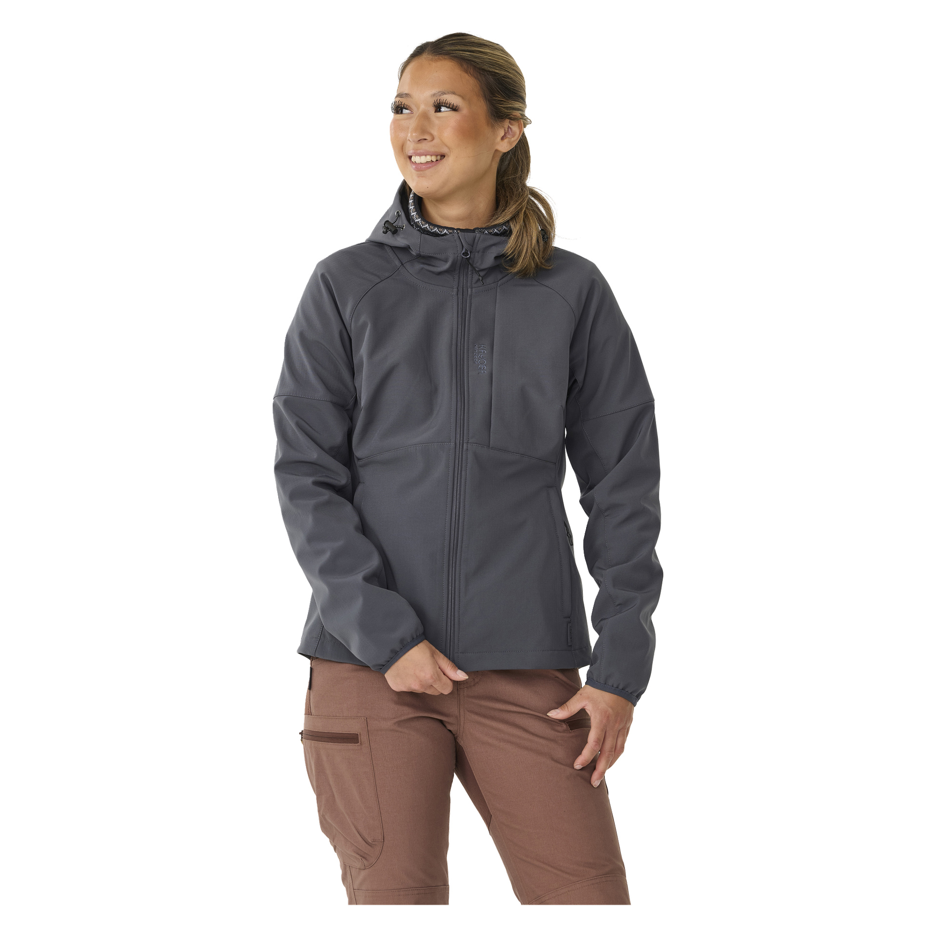 Eine lächelnde Person trägt die graue Kayoba Outdoor Damenjacke Hede mit Fleecefutter gefüttert Softshell mit Reißverschluss vorne, Seitentaschen, Kapuze mit verstellbaren Kordeln und brauner Hose. Ihr Haar ist zu einem Pferdeschwanz gestylt.