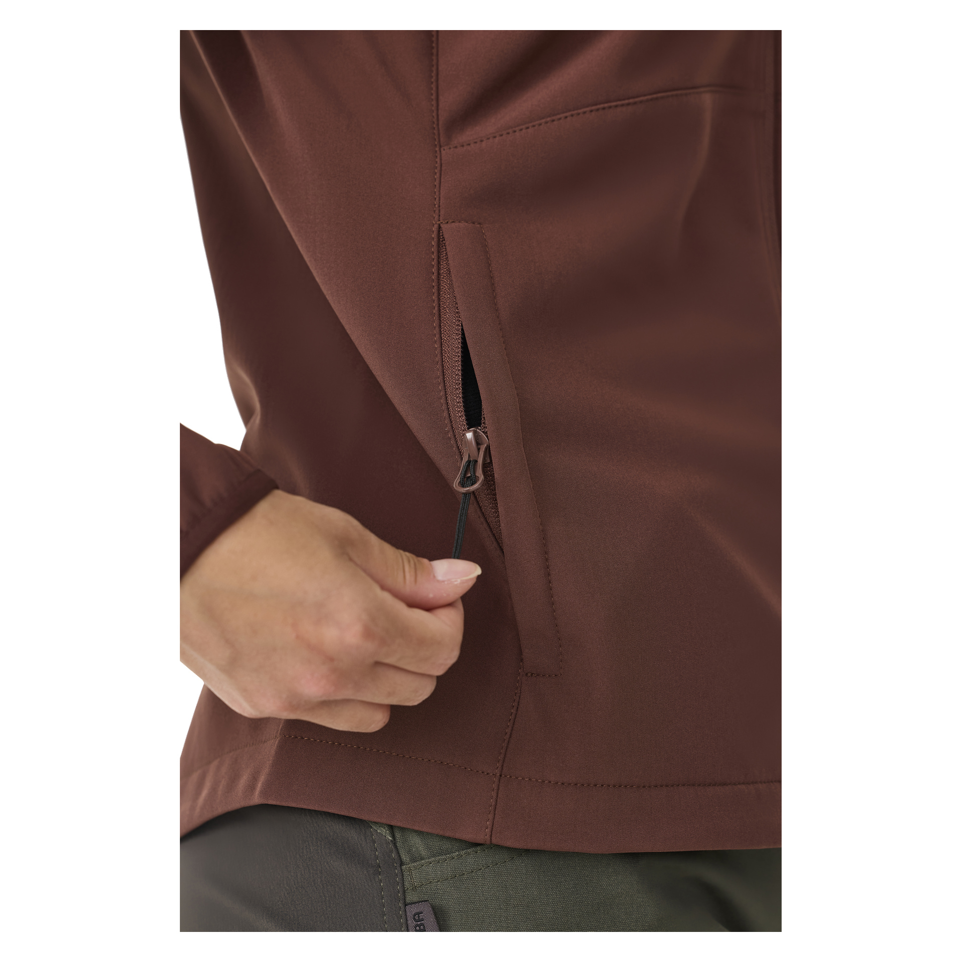 Eine Hand zieht den Reißverschluss an der Seitentasche einer braunen Kayoba Outdoor Women's Softshell-Softshelljacke Hede, die mit grünen Hosen am unteren Rand des Bildes teilweise sichtbar ist und sich auf den Taschenbereich konzentriert.