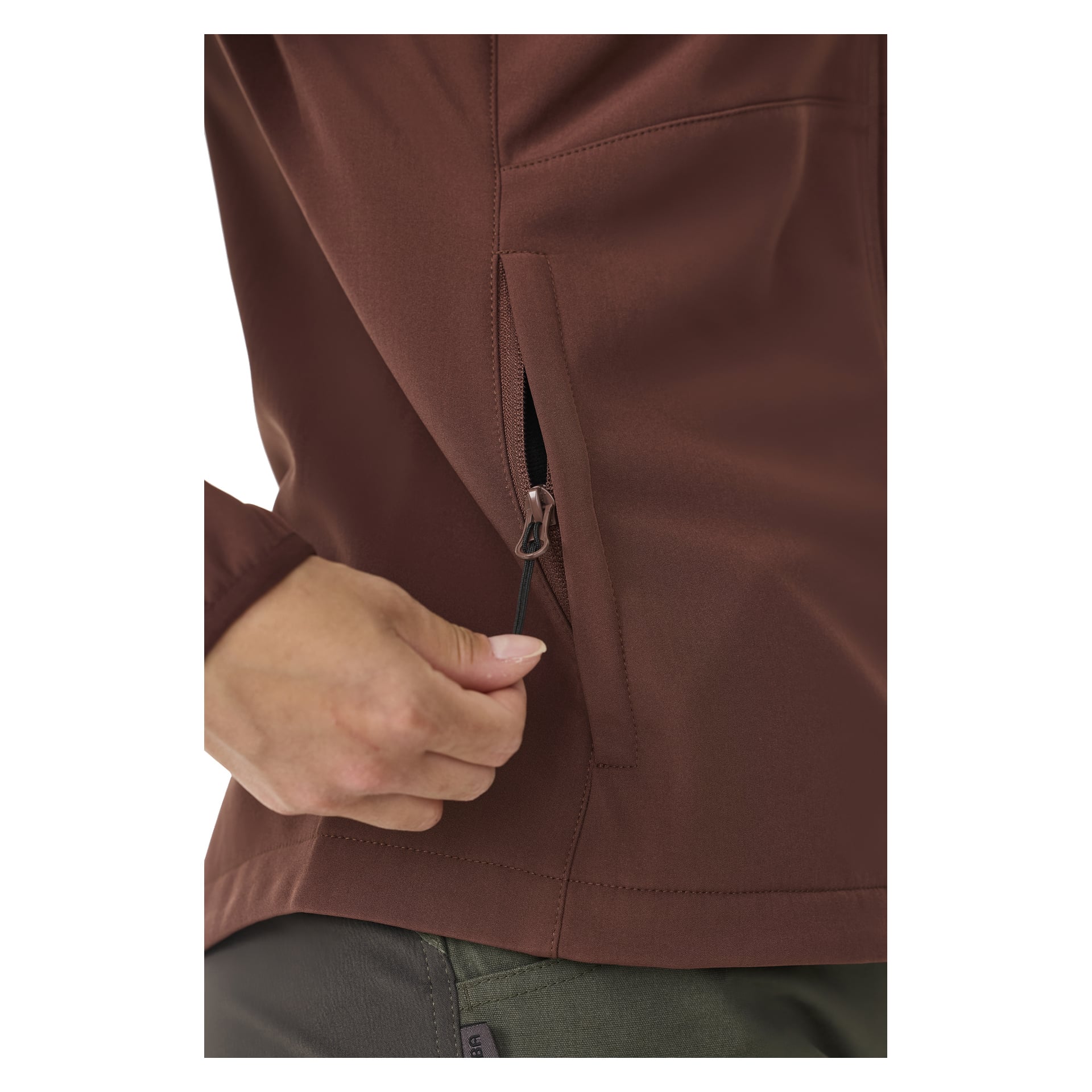 En hand drar i dragkedjan på sidofickan på en brun Kayoba Outdoor Women's fleecefodrade softshell-jacka Hede, med gröna byxor delvis synliga längst ner på bilden, med fokus på fickområdet.