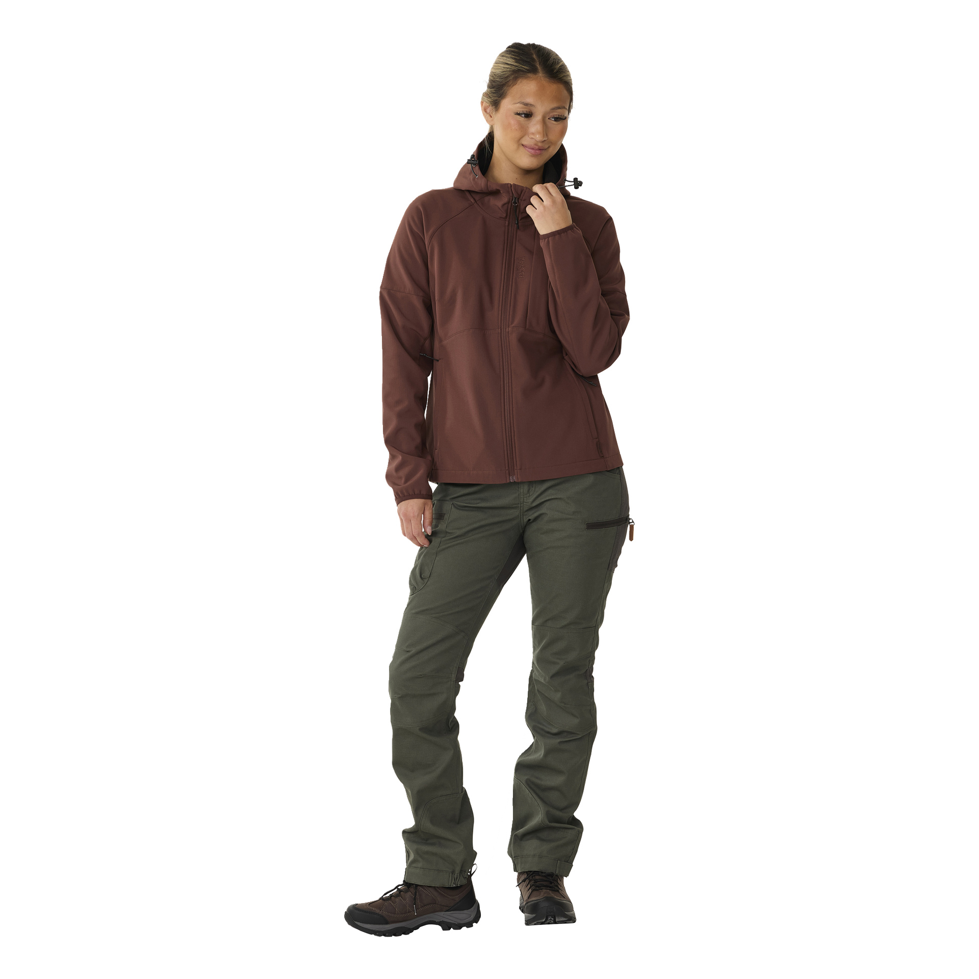 Eine Person ist leicht zur Seite gedreht und trägt eine kastanienbraune Kayoba Outdoor Outdoor Softshell-Softshell-Jacke Hede, olivgrüne Outdoor-Hosen und schwarze Wanderschuhe. Eine Hand ruht am Kinn und ihr Gesichtsausdruck ist neutral.