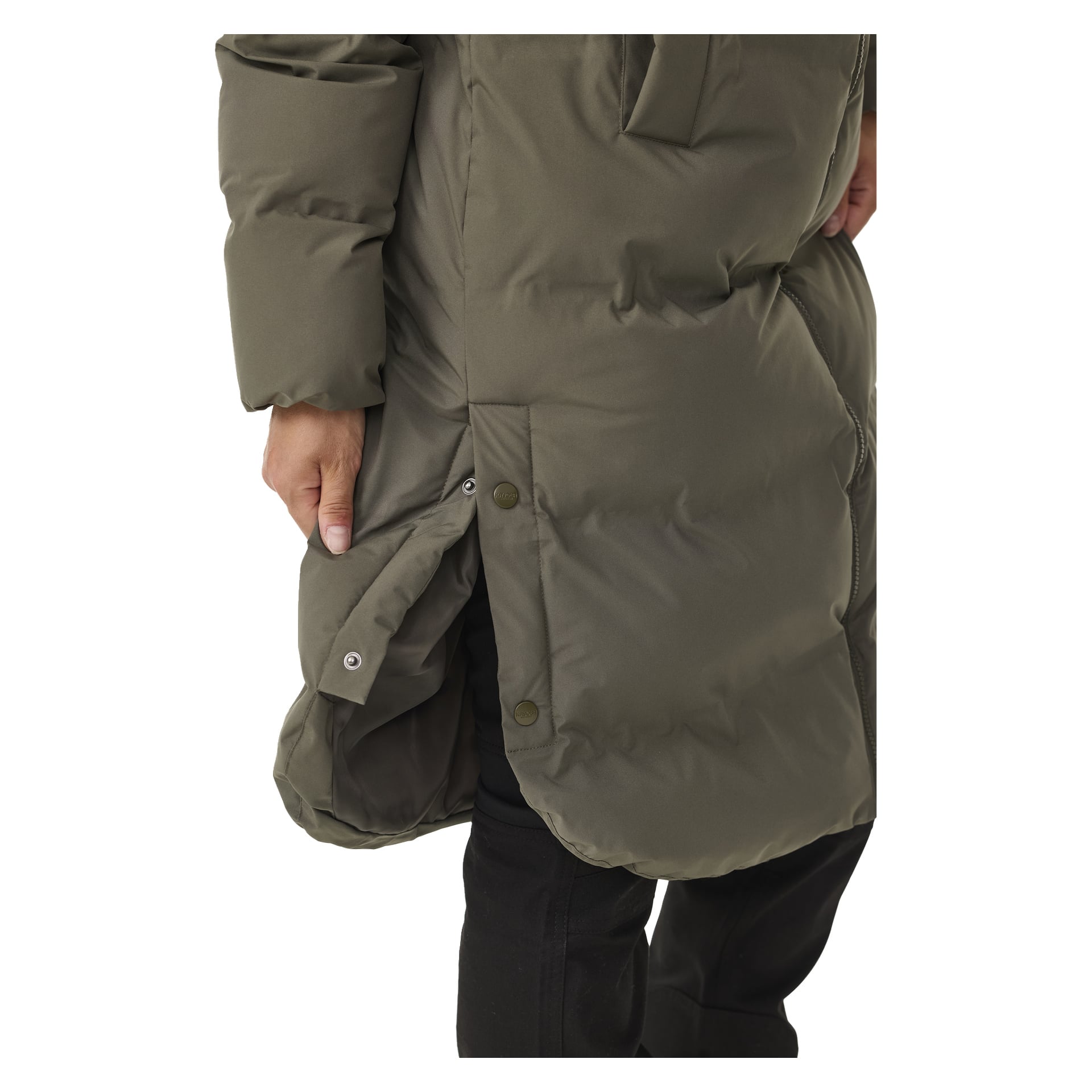 En närbild visar en person klädd i den olivgröna Kayoba Outdoor Women's vinterkappa Storlien, med knappknappar längs sidan. Kappan är delvis knäppt, vilket blottar svarta byxor under, och deras hand är placerad nära jackans öppning.