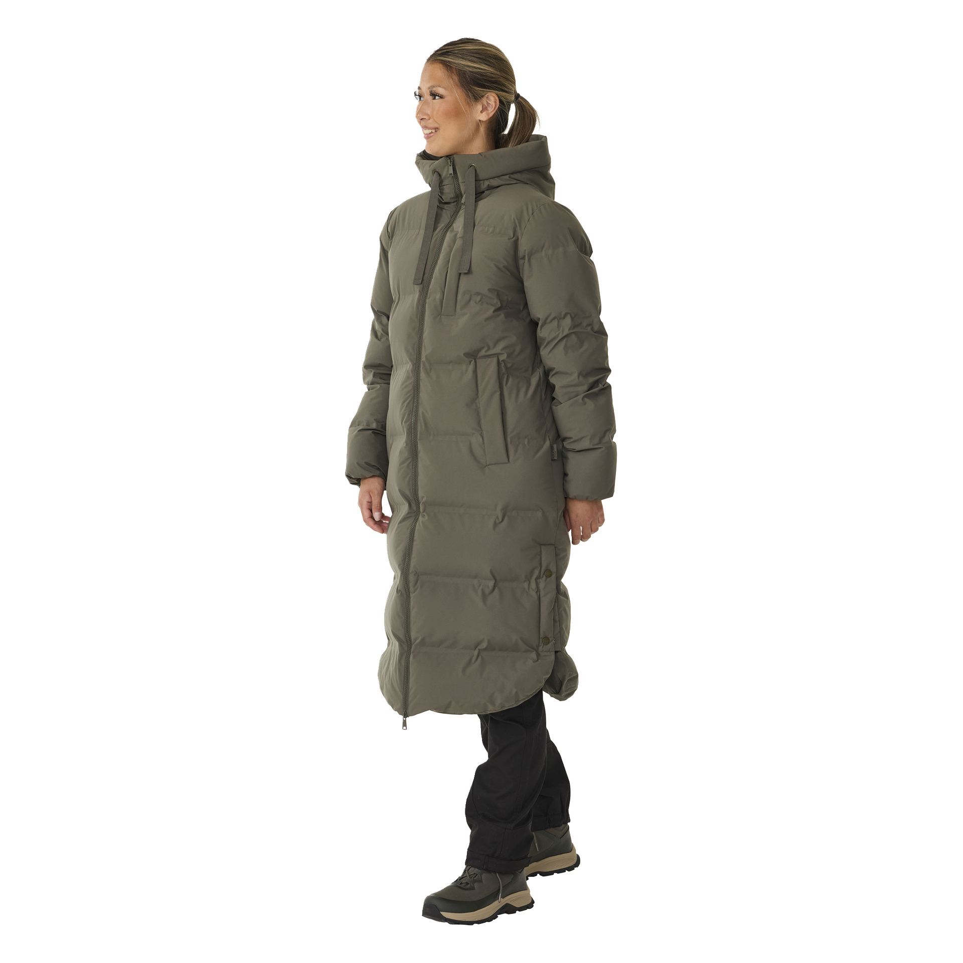 Henkilö mallintaa Kayoba Outdoor Women's Storlien -talvitakkia, pitkää oliivinvihreää hupparia, jossa on suuret taskut, vetoketju kiinni ja käytetään mustien housujen ja polkukenkien kanssa. Heidät on kuvattu profiilissa vasemmalle päin, hymyillen hieman.