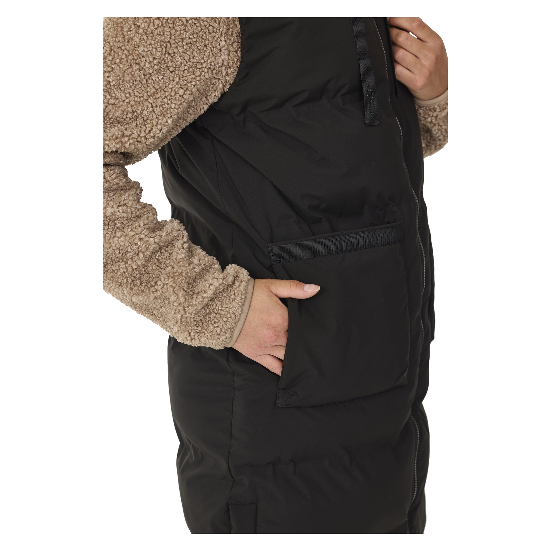 Henkilö, jolla on vaaleanruskea fleece-hiha ja musta Kayoba Outdoor Pehmustettu naisten lilet, Long Duved, on osittain piilottanut oikean kätensä suureen etutaskuun. Kuva on rajattu niin, että paljastuu vain heidän vartalonsa ja osan käsivarrestaan.