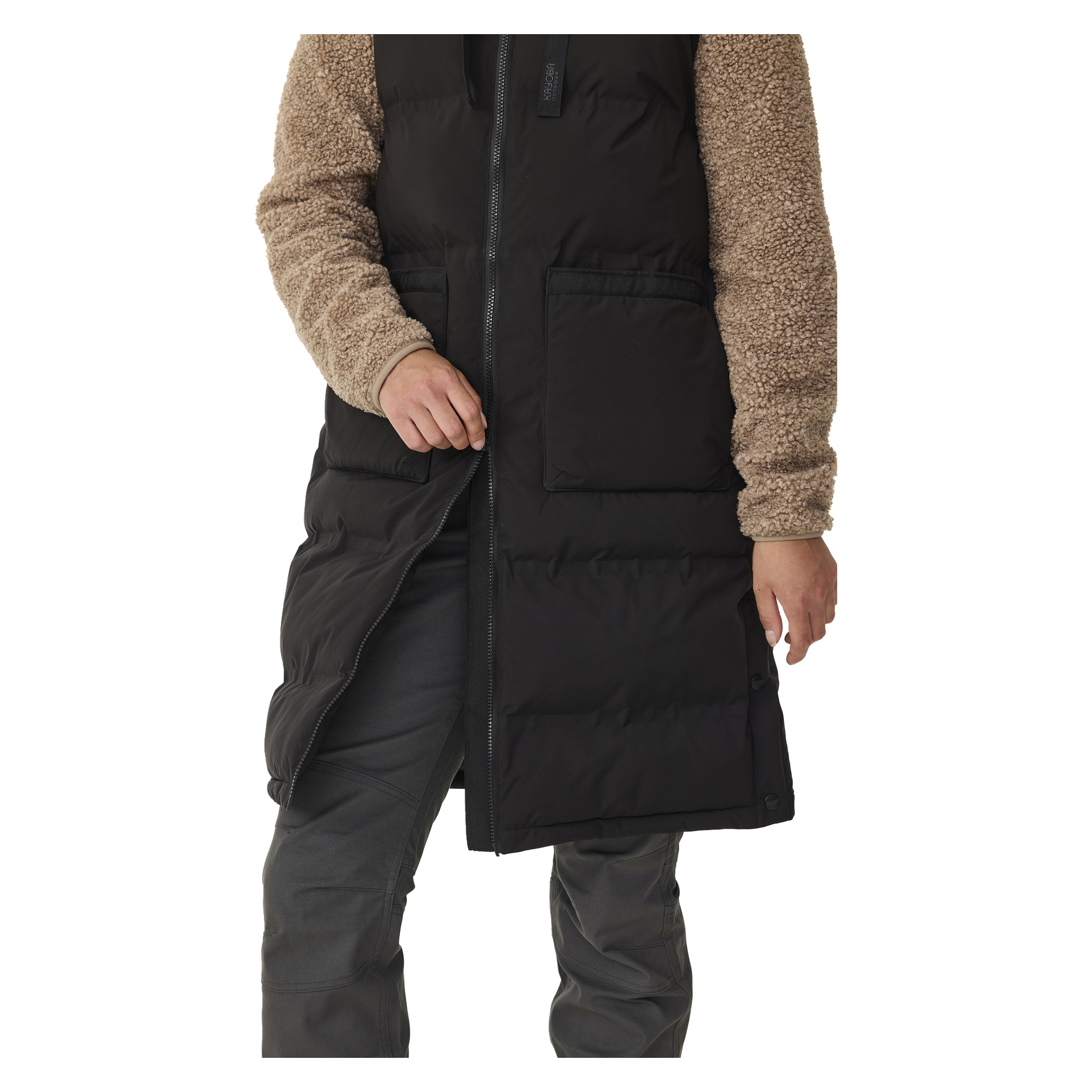 Man öffnet den langen Damen-Gilet von Kayoba Outdoor Padded, der über einer beigefarbenen Fleece-Ärmeljacke laget. Das Bild zeigt nur den Unterkörper mit grauen Hosen.