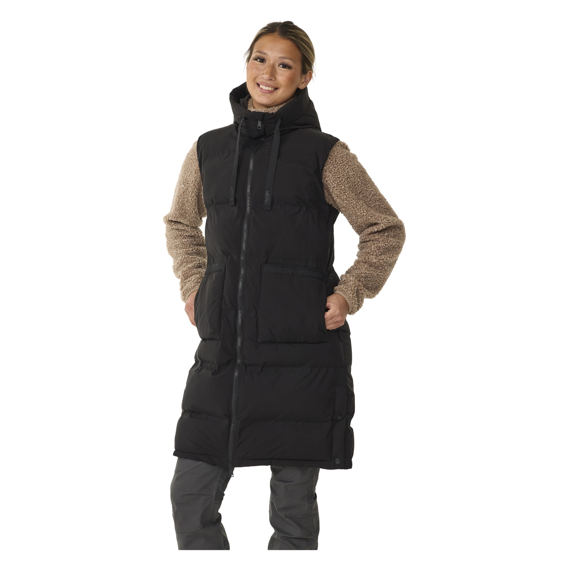 En leende person visar Kayoba Outdoor Padded Women's Gilet, Long Duved – en svart pufferväst med framträdande framfickor, huva och dragkedja. De har på sig en beige långärmad skjorta under och grå byxor, med båda händerna i västfickorna.