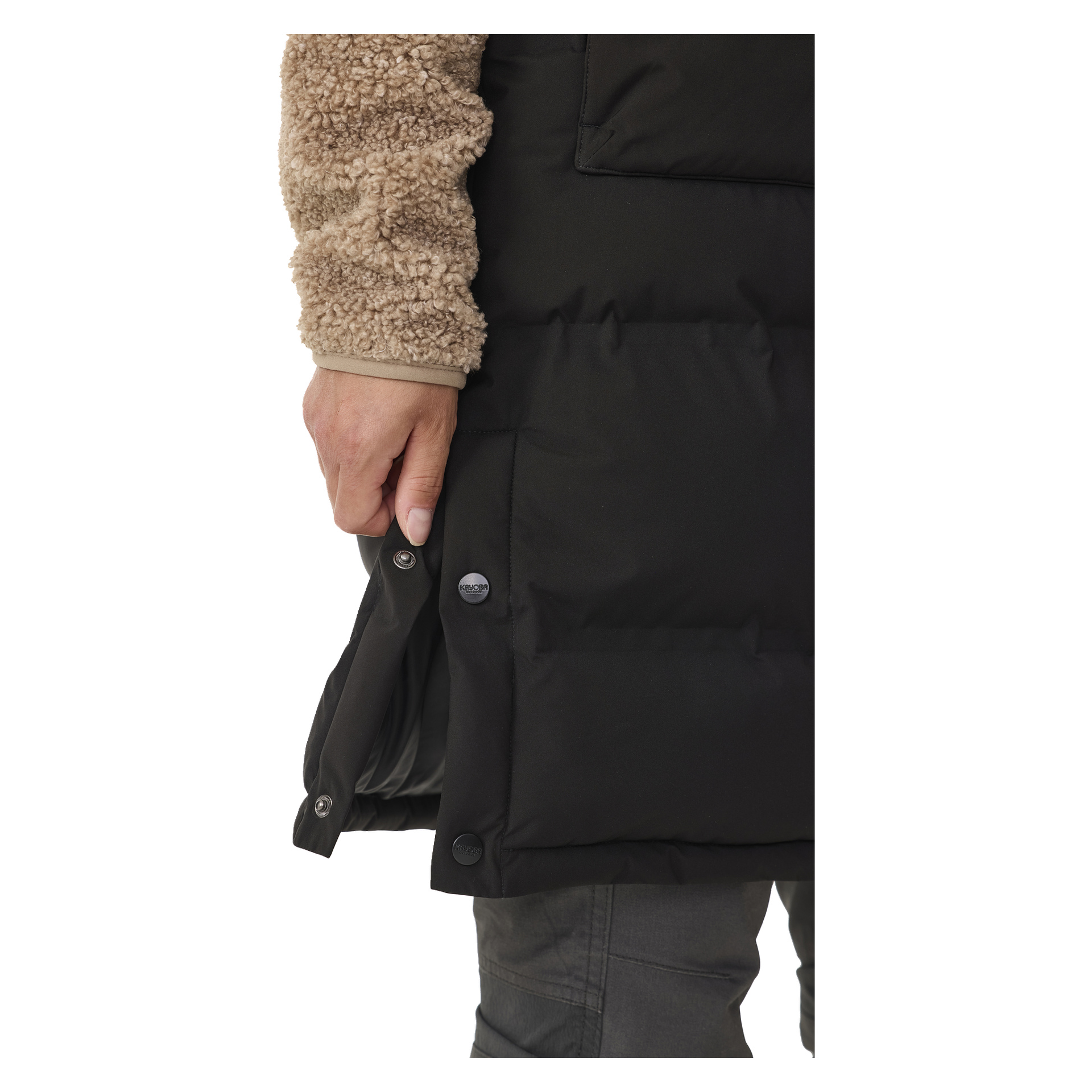 Henkilö, joka on pukeutunut beige-fleece-takkiin ja harmaisiin housuihin, on kuvattu kiinnittämässä nappeja Kayoba Outdoor Padded Women's Gilet, Long Duved, sivussa, kiinnittäen huomiota liivin sivuavaukseen.