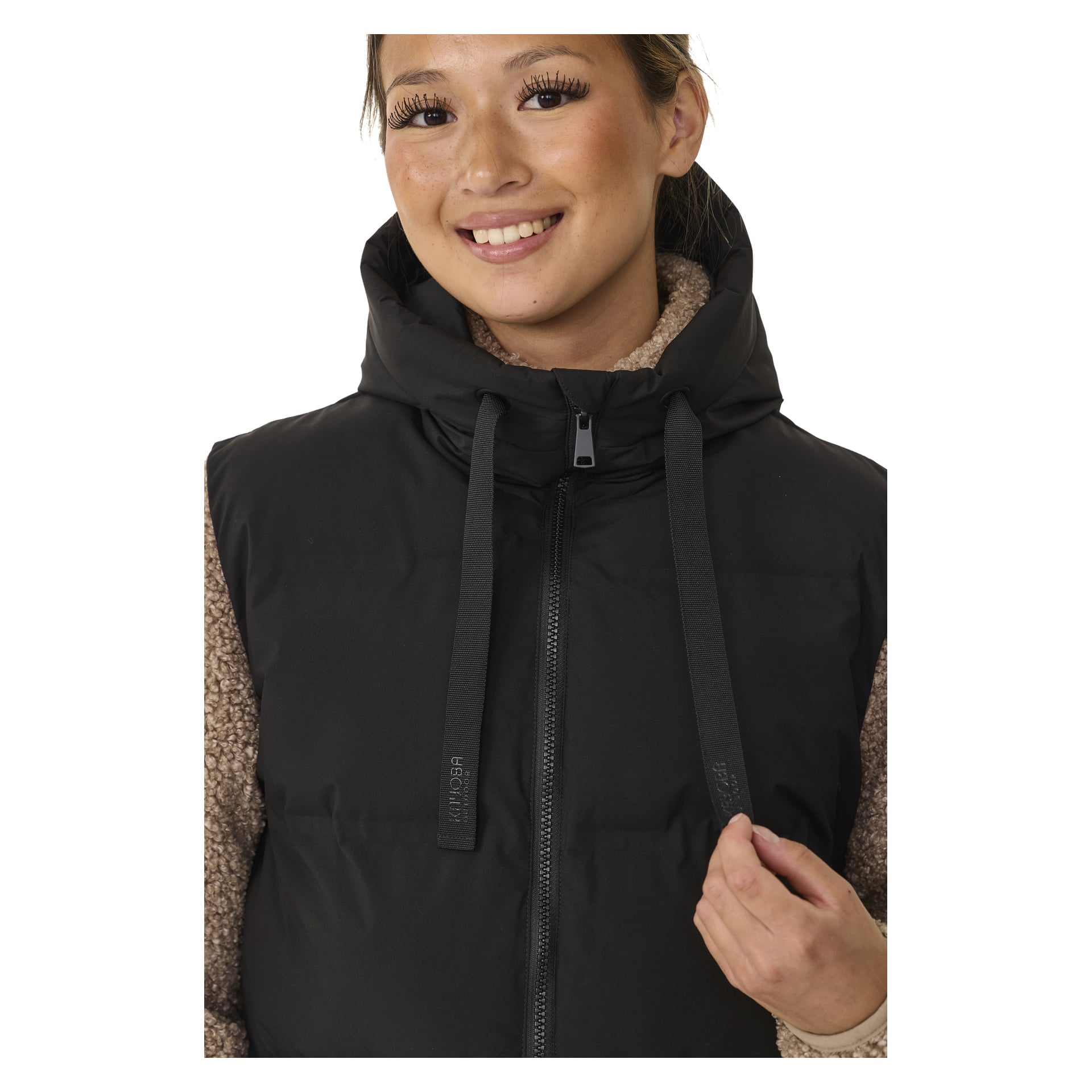 Henkilö hymyilee kameralle, pukeutuneena mustaan Kayoba Outdoor Pehmustettuun Naisten Gilet-paitaan, Long Duved, kerroksittain vaaleanruskean neuleen päällä. Liletissä on huppu, etuvetoketju ja pitkät nyörit.