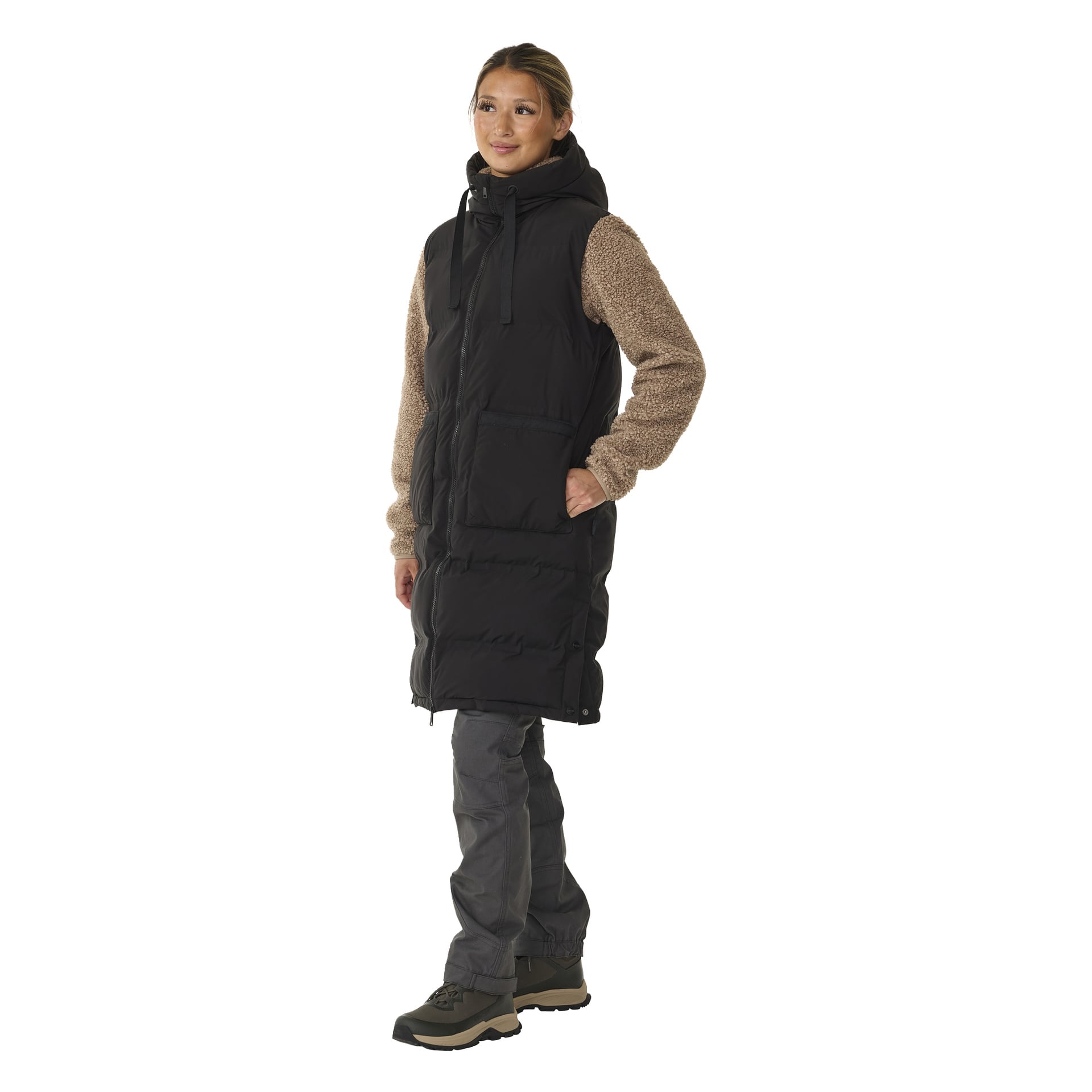 Nainen mallintaa pitkän Kayoba Outdoor Padded -naisten giletin vaaleanruskean neuleen päällä, harmaisiin housuihin ja ruskeisiin saappaisiin. Hän seisoo vinossa, toinen käsi lilet-taskussa, pitäen neutraalin ilmeen.