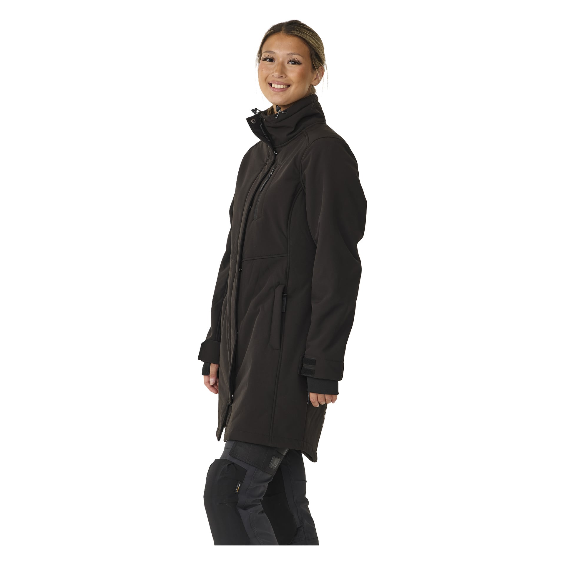 Eine Frau präsentiert die Bluewear Woman's Softshell Jacket, Long Gunnel – einen eleganten, dunklen Mantel mit hohem Kragen und Reißverschlusstaschen. Sie trägt dunkle Hosen und lächelt sanft, während sie zur Seite schaut.