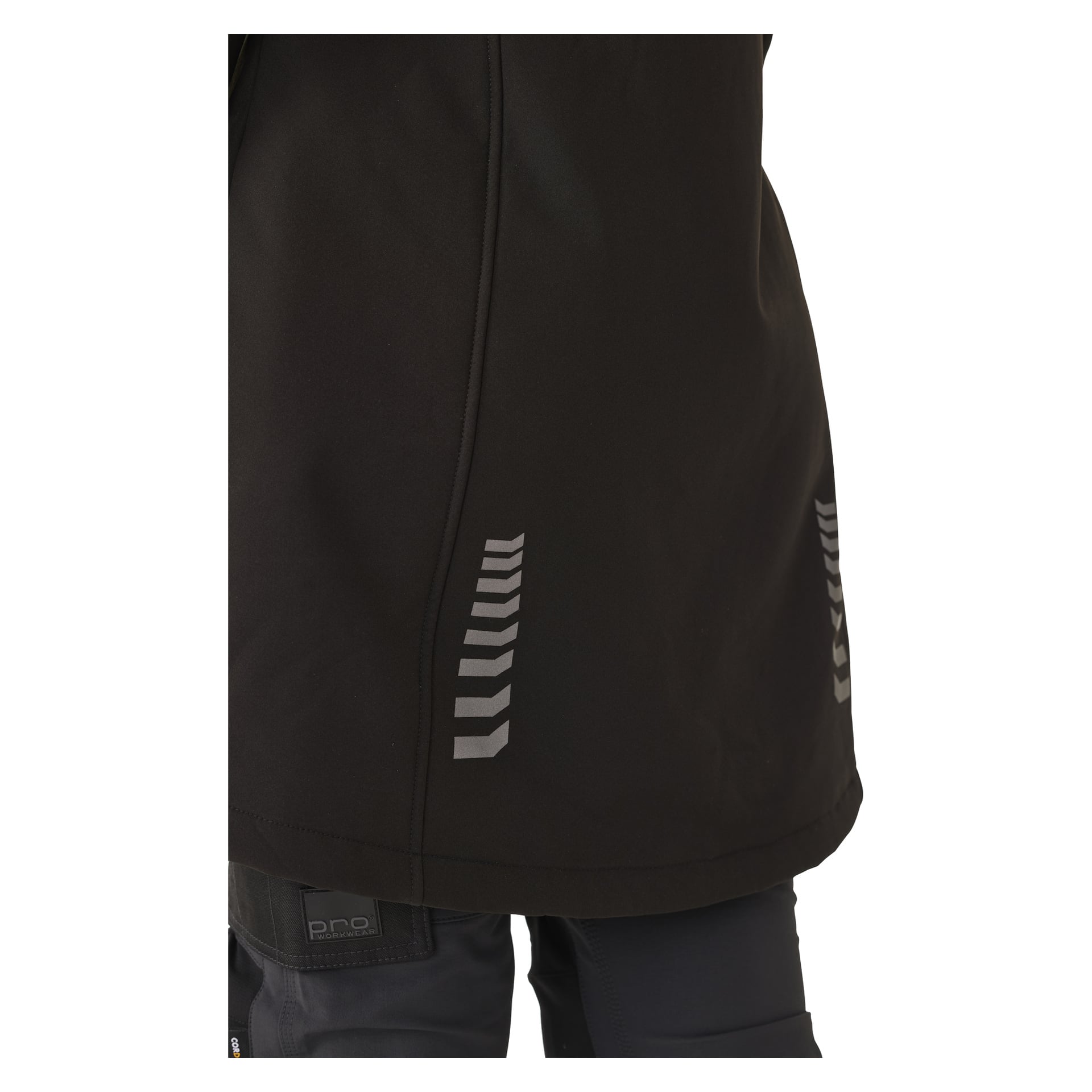Eine Nahaufnahme zeigt eine Person in der langen Gunnel-Softshell-Jacke der Bluewear Woman in Schwarz, mit mehreren kleinen reflektierenden Chevron-Details nahe dem Saum an der Seite. Das Bild zeigt den unteren Teil der Jacke zusammen mit einem Teil der oberen Hose.