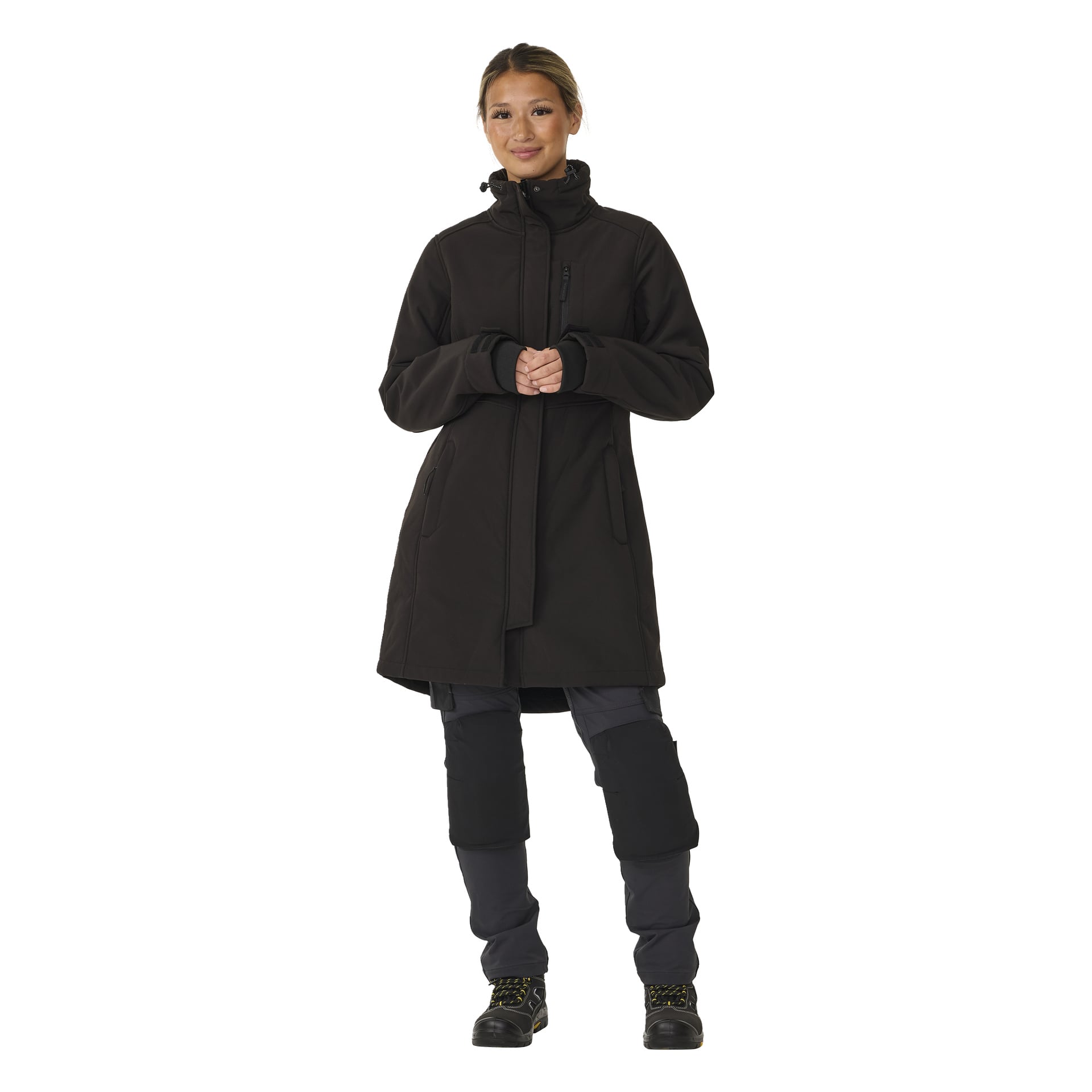 Eine Person schaut nach vorne, gekleidet in die lange Softshell-Jacke der Bluewear Woman namens Gunnel, dazu dunkle Hosen mit Knieschonern und schwarze Stiefel mit gelben Akzenten. Ihre Hände sind vor ihnen verschränkt.