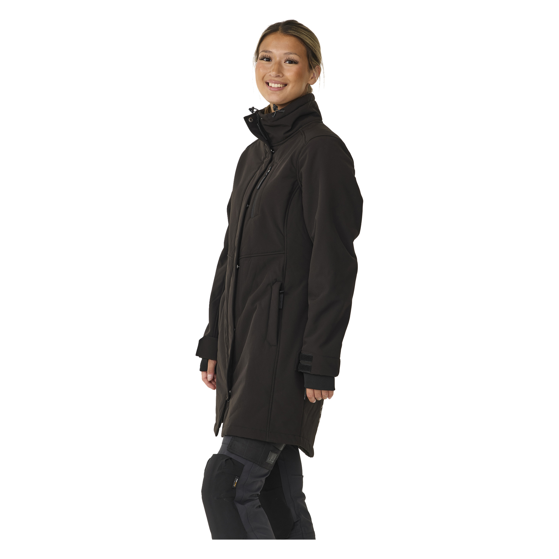 Henkilö seisoo hieman vinossa ja hymyilee, mallintaen Bluewear Womanin pitkää Gunnel-softshell-takkia – mustaa takkia, jossa on vetoketju, korkea kaulus ja taskut.