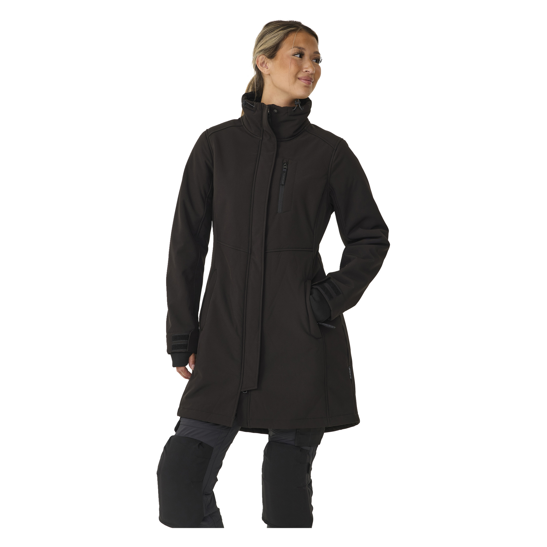 Henkilö mallintaa Bluewear Women's long Gunnel softshell -takin, mustan vetoketjullisen takin, jossa on korkea kaulus. Toinen käsi on taskussa, he katsovat sivulle ja hiukset on vedetty matalalle poninhännälle.