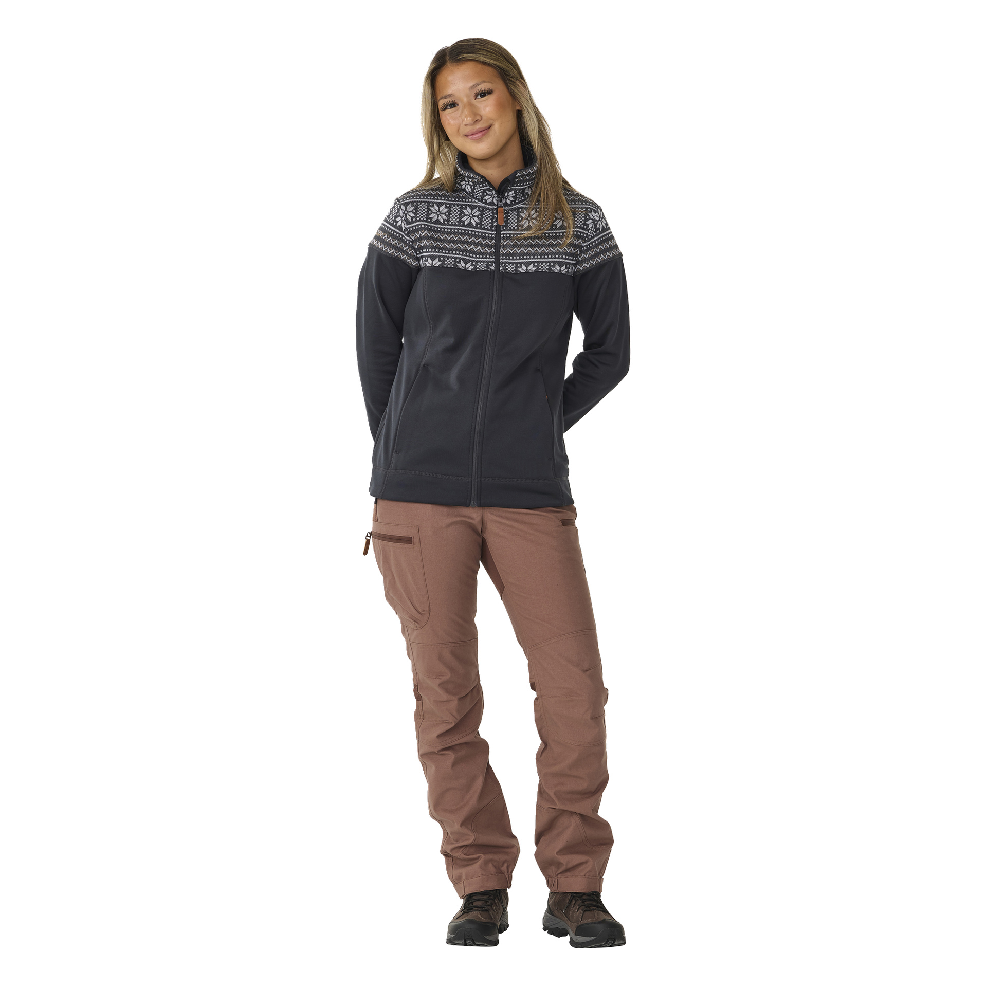 Henkilö, jolla on pitkät hiukset ja lempeä hymy, on kuvattu eteenpäin, pukeutuneena Kayoba Outdoor Women's mid-layer -neuleeseen Valbo, jonka yläosassa on valkoinen geometrinen kuvio. Heillä on myös vaaleanruskeat ulkohousut ja mustat kengät.