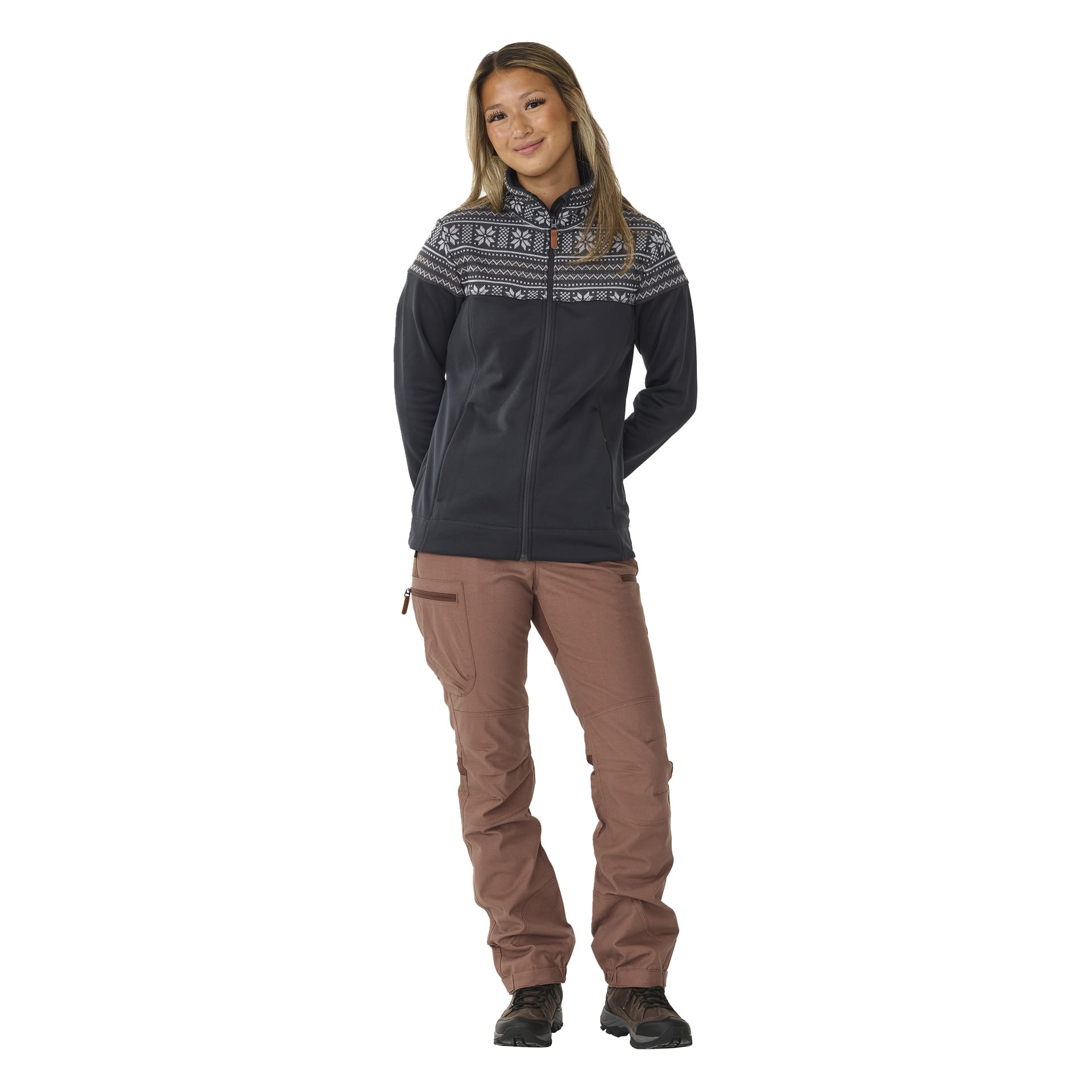 En person med långt hår står framåtvänd med händerna i fickorna, klädd i Kayoba Outdoor Women's Valbo mellanlagertröja. Tröjan har svart dragkedja framtill och ett vitt geometriskt mönster över bröstet. De bär också ljusbruna byxor med sidofickor och svarta skor.