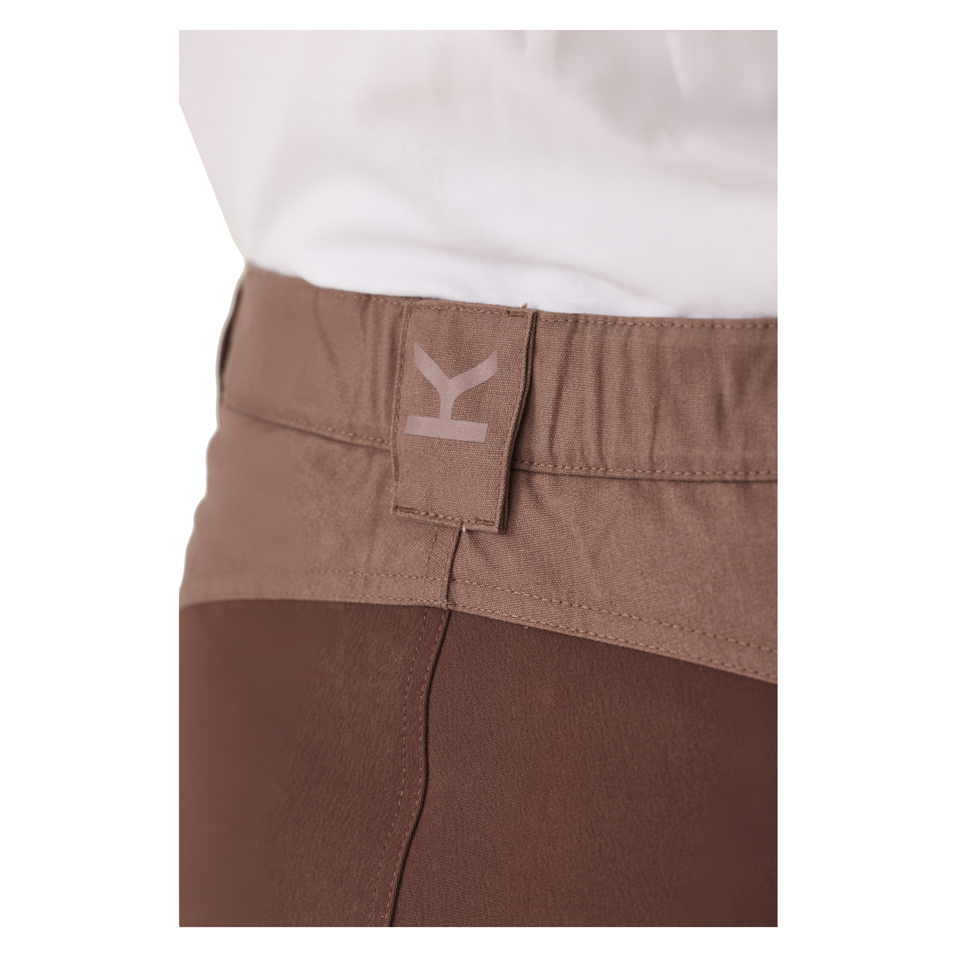 Detaljert visning av den brune midjestrikken på damebuksene Älvdalen 2.0 Leisure fra Kayoba Outdoor, med en beige og en stilisert Y-logo. En hvit skjorte er pent stukket ned i buksa.