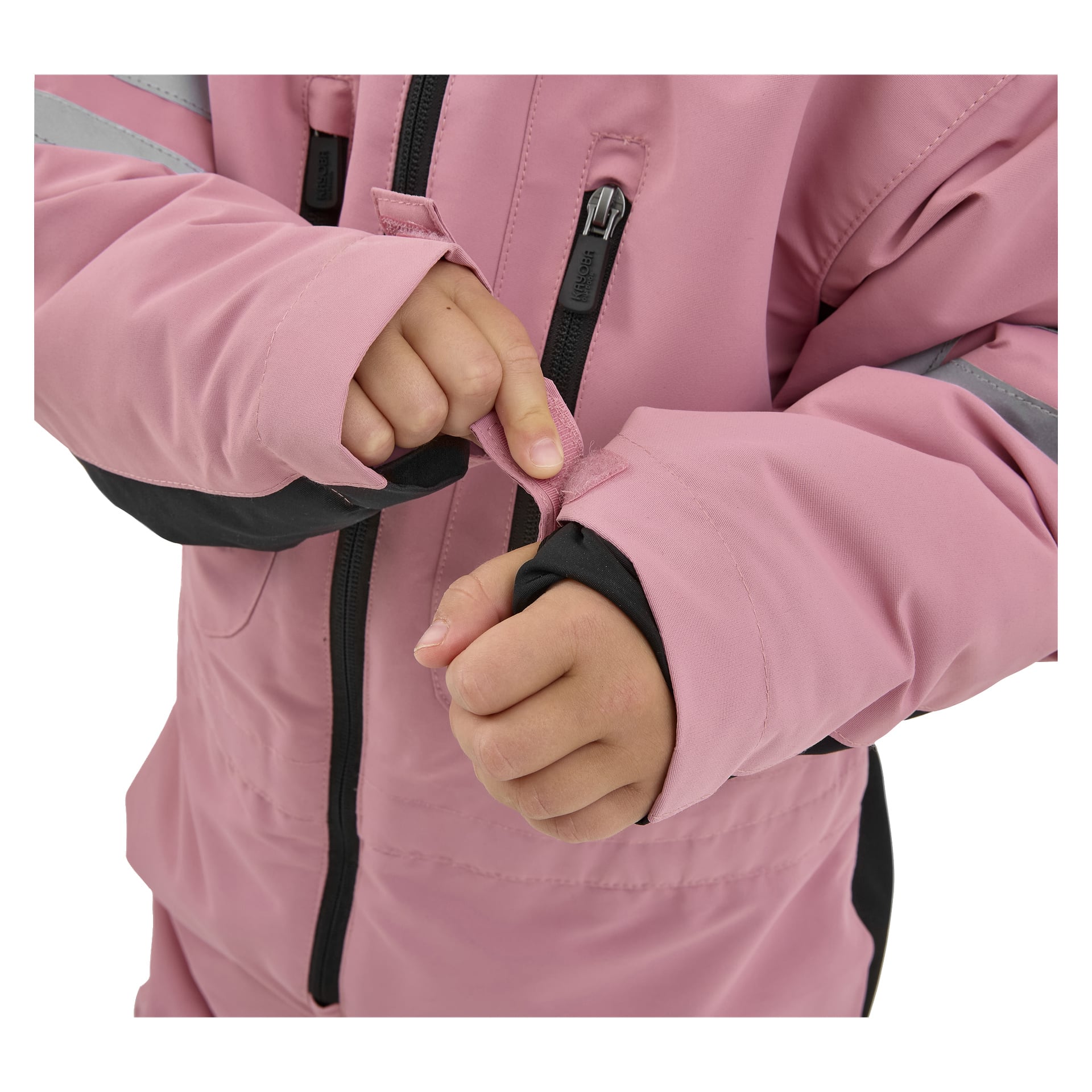 Ett barn justerar den krok-och-ögla remmen på den rosa ärmen på en Dundret-jacka från Kayoba Outdoor, som har svarta dragkedjor och en reflekterande armremsa. Bilden visar bara barnets händer och en del av jackan.